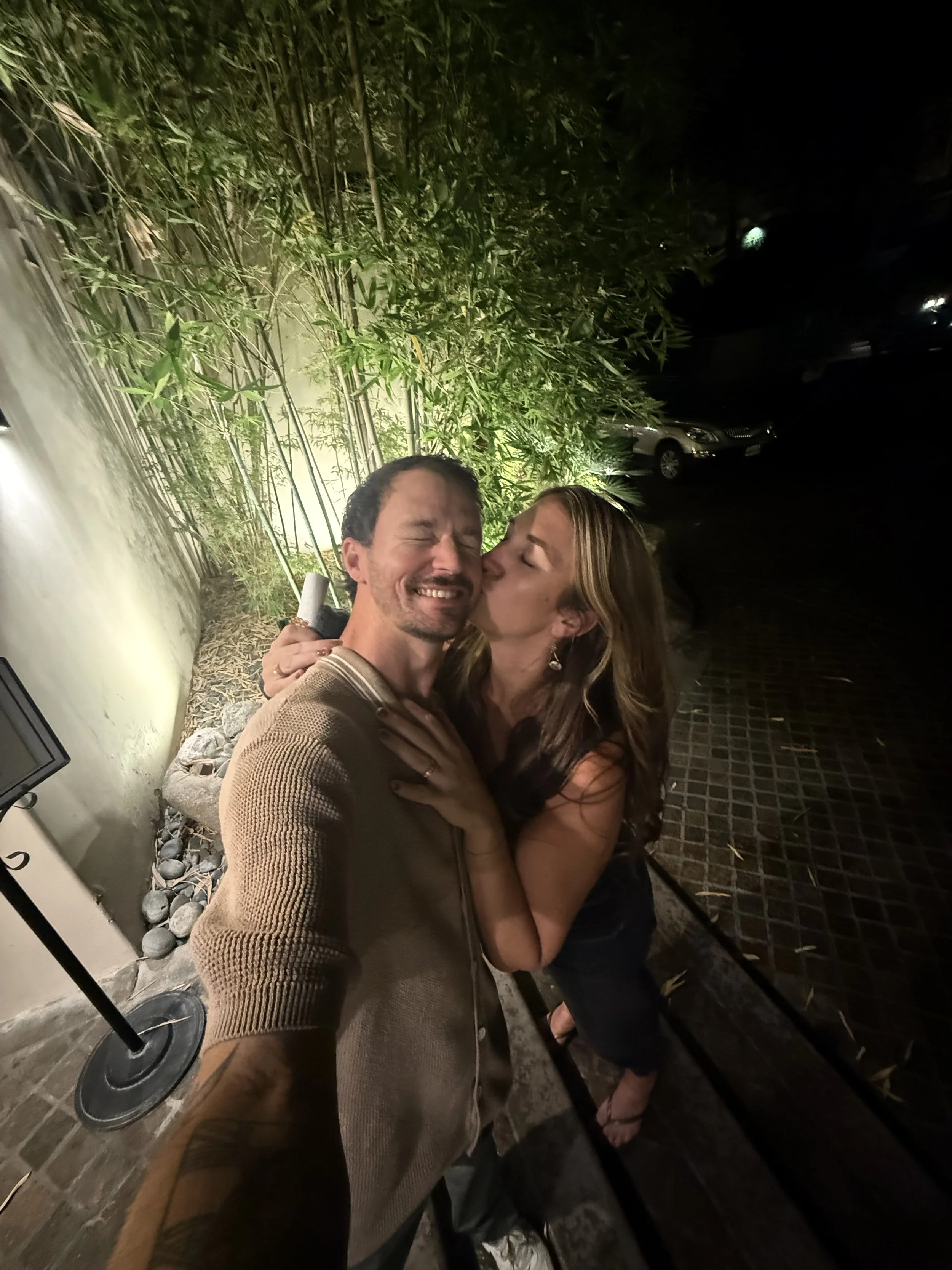 A man and a woman sharing a kiss, with the woman holding a cigarette, standing outdoors at night near a bamboo garden and parked cars.