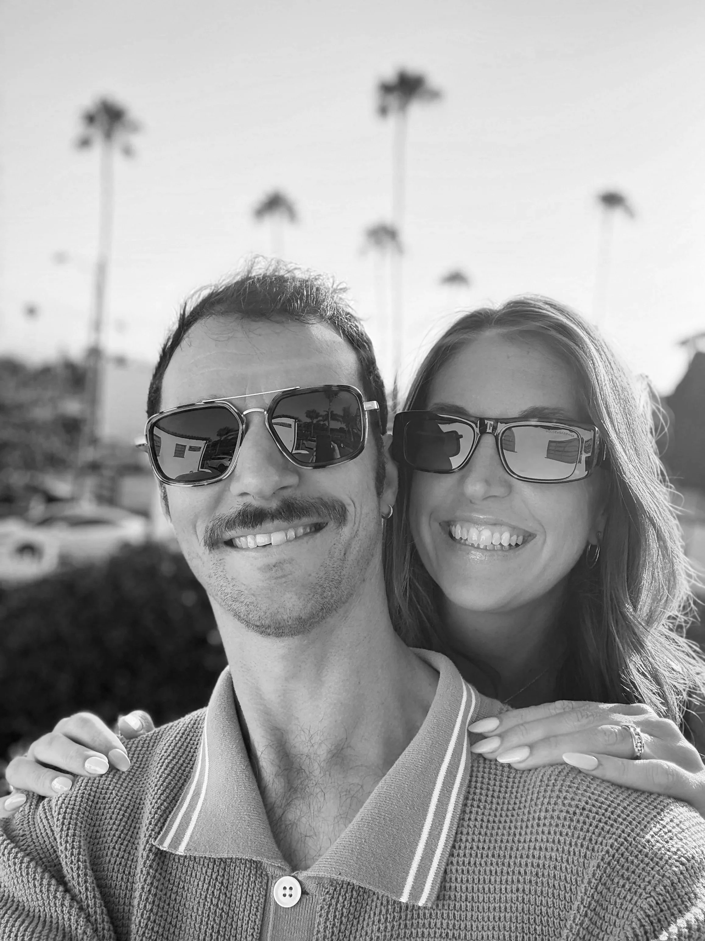 A black and white photo of a smiling man and woman wearing sunglasses, standing outdoors with palm trees in the background.