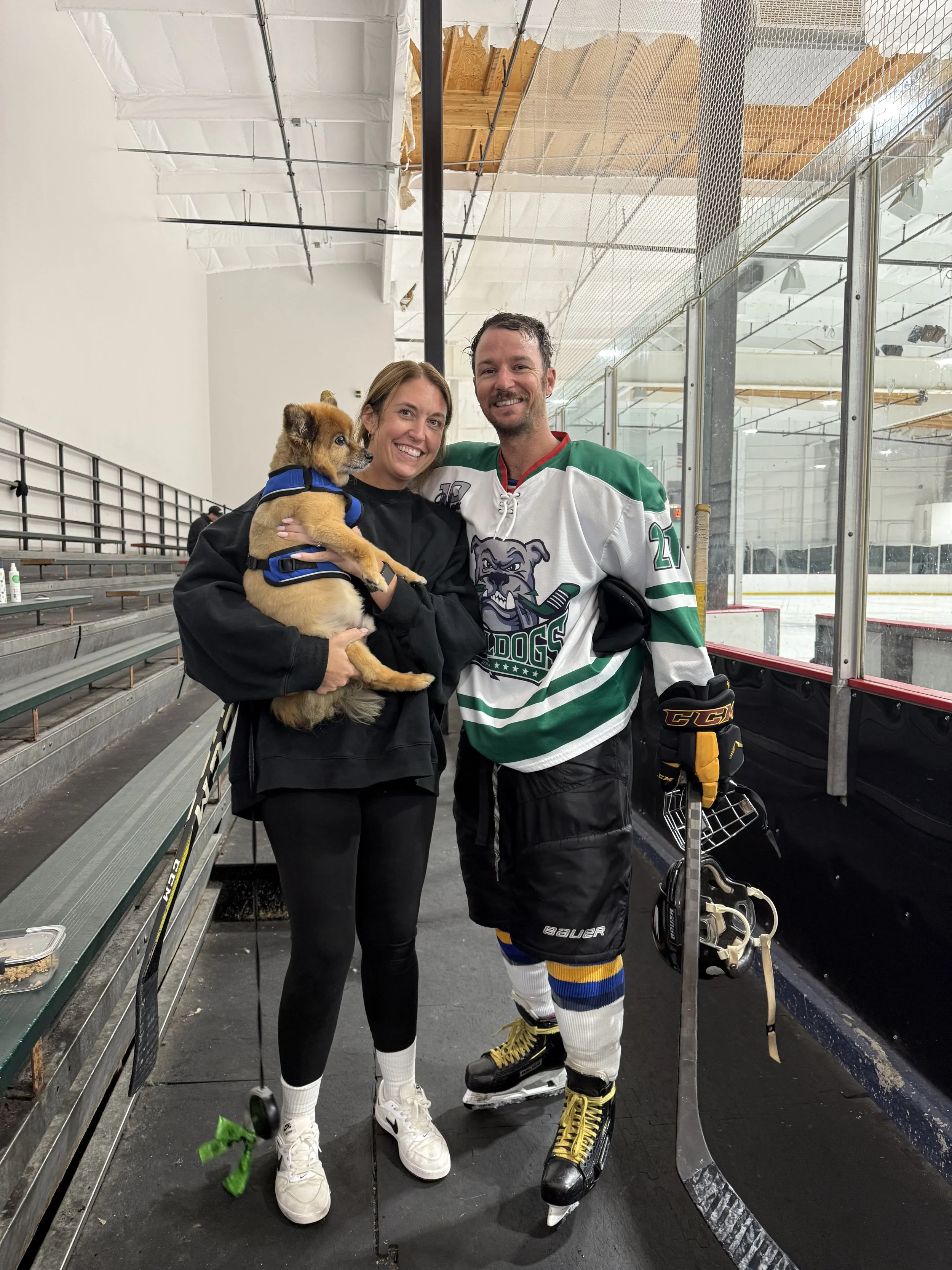 A woman and a man, both smiling, standing on an ice rink. The woman is holding a small dog. The man is wearing a hockey uniform, including skates and holding a hockey stick.
