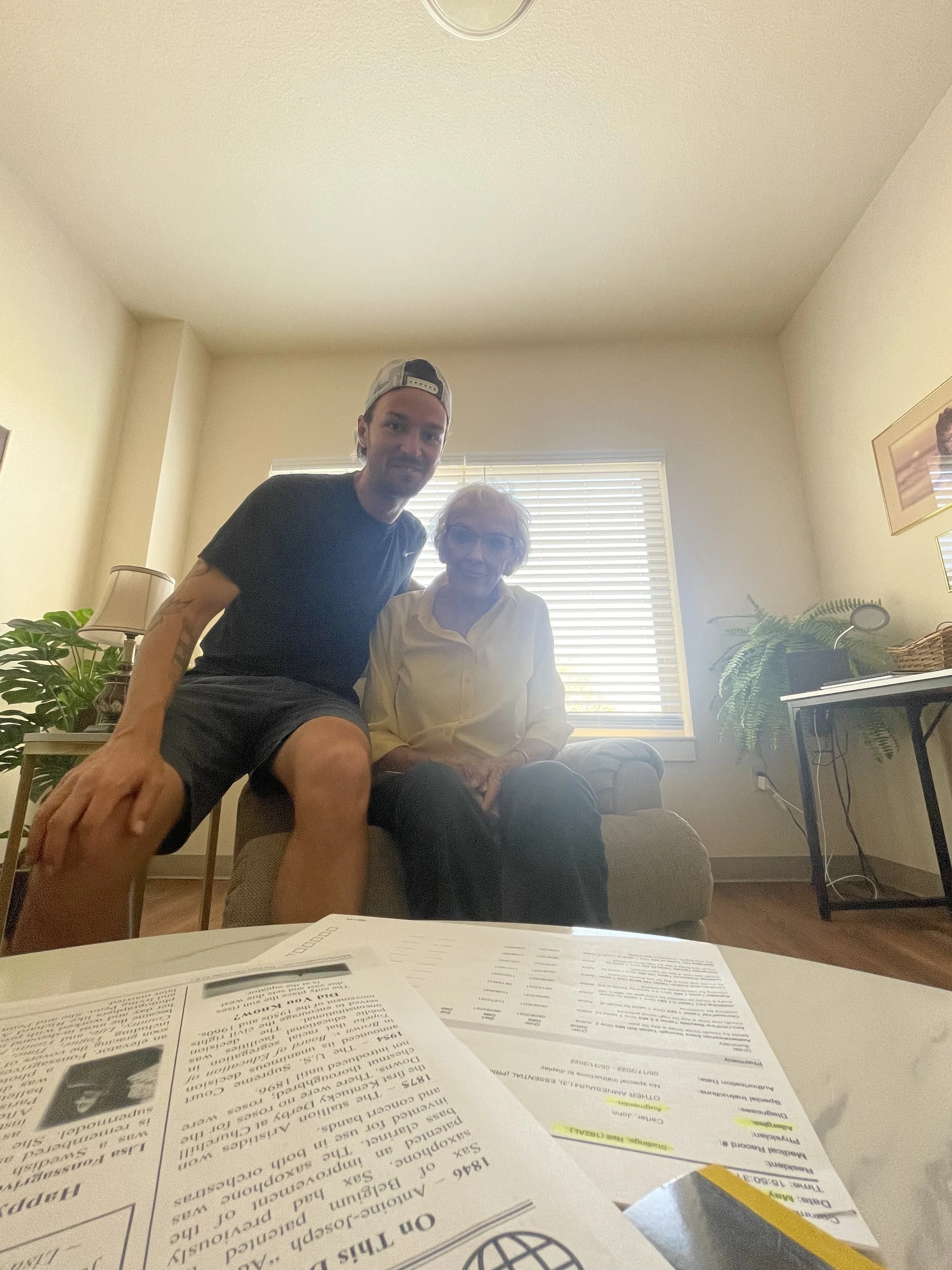 A man and an elderly woman sitting together in a living room with blinds on the window behind them, plants on side tables, and papers or documents on a table in front of the camera.
