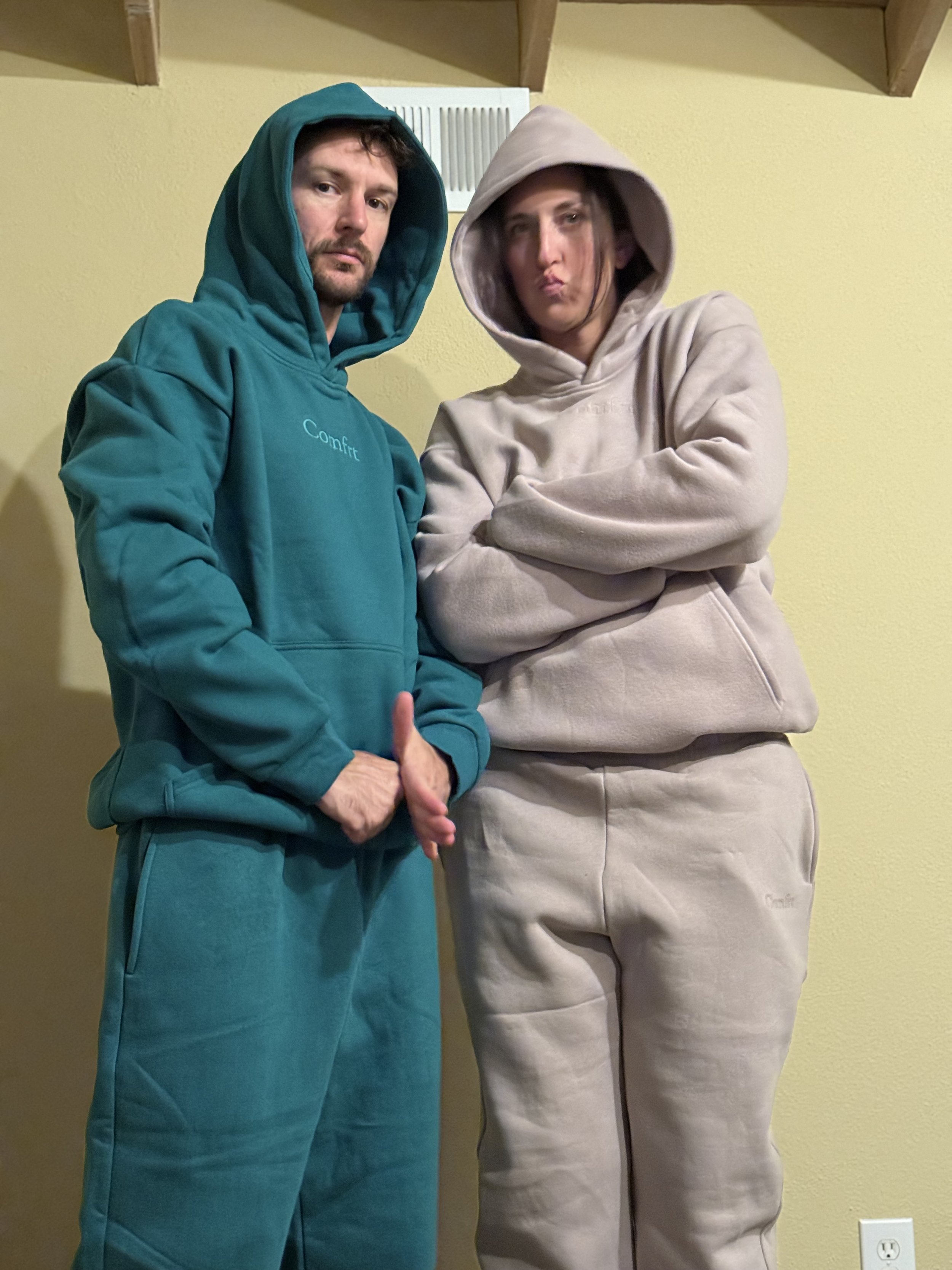 Two people standing against a yellow wall, both wearing hooded sweatshirts. The man on the left wears a teal hoodie and pants with his hands clasped in front of him. The woman on the right wears a beige hoodie and pants, with her arms crossed and lip