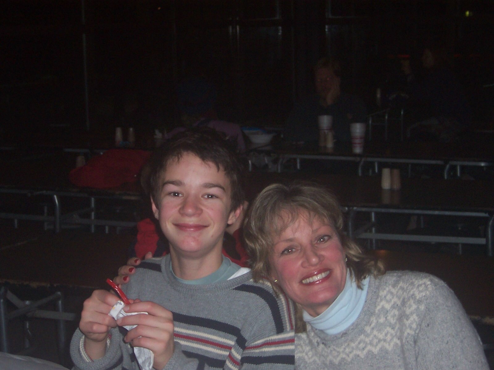A woman with curly hair and a light-colored sweater smiles with her arm around a young man with short brown hair in a striped sweater. They are in a dimly lit room with empty tables and cups in the background.