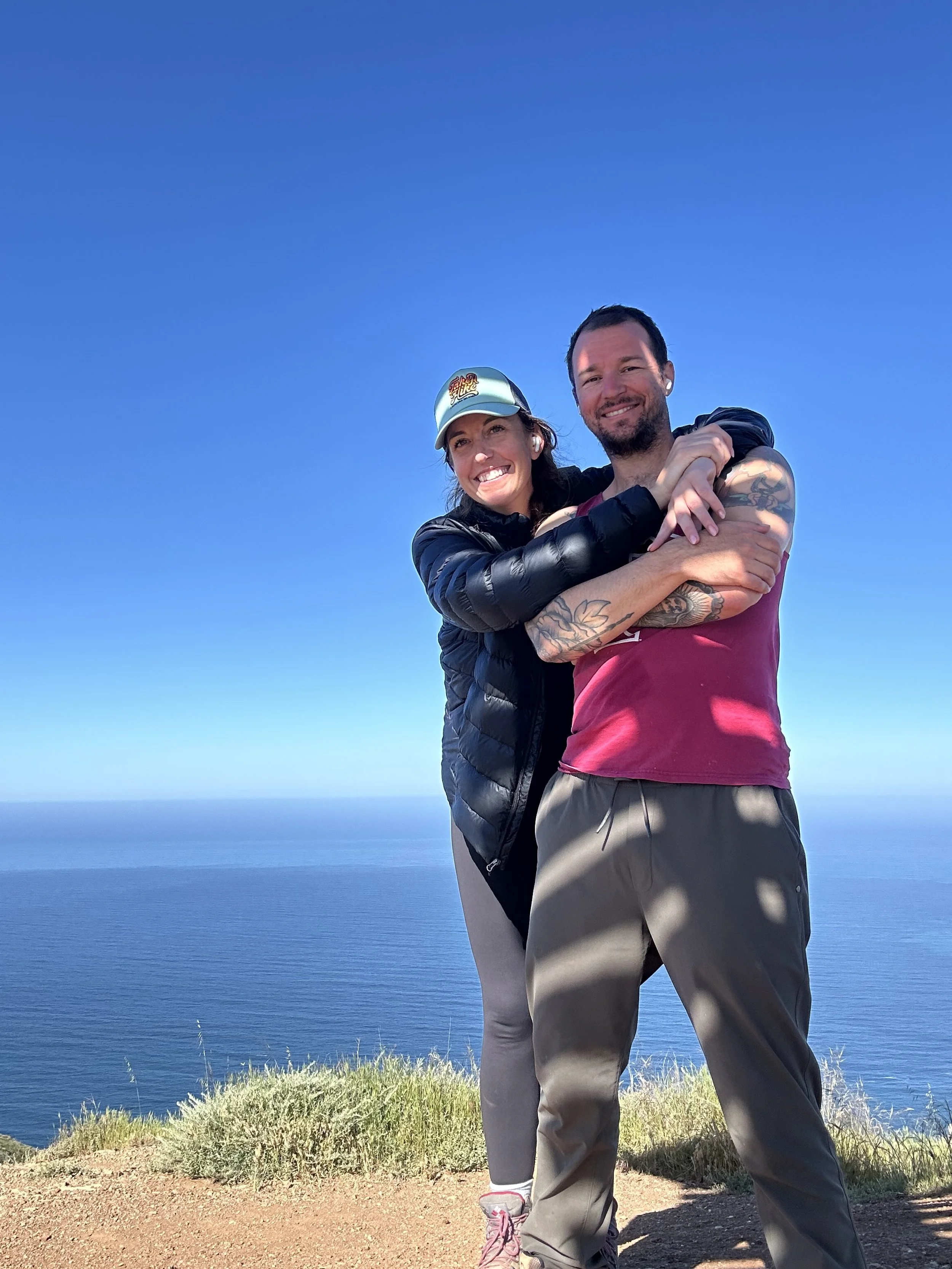 A smiling couple hugging outdoors on a sunny day with a view of the ocean and clear blue sky in the background.