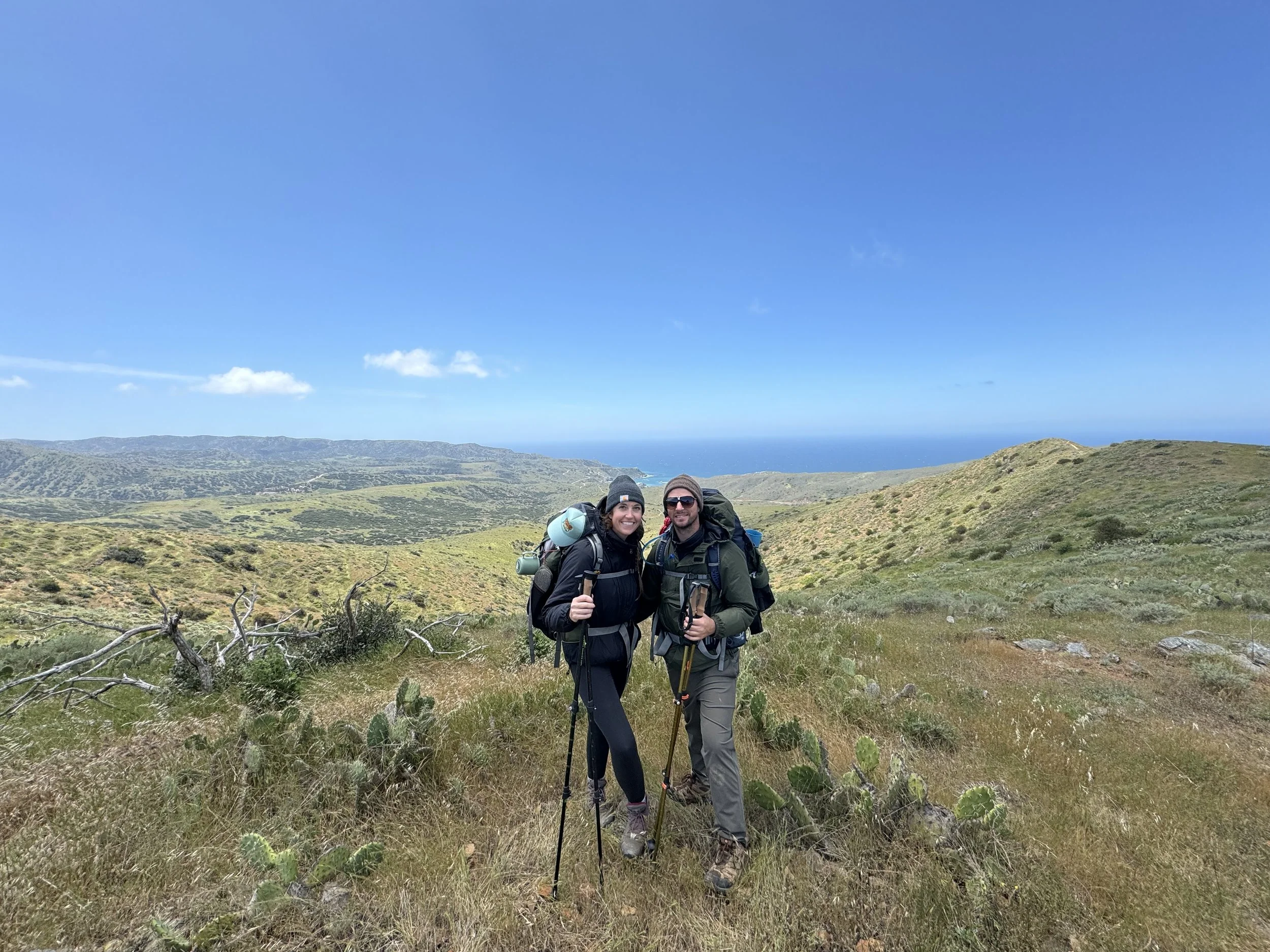 Two hikers, a woman and a man, standing on a grassy trail in a hilly landscape with green vegetation, cacti, and shrubs, with the ocean visible in the background under a blue sky with a few clouds.
