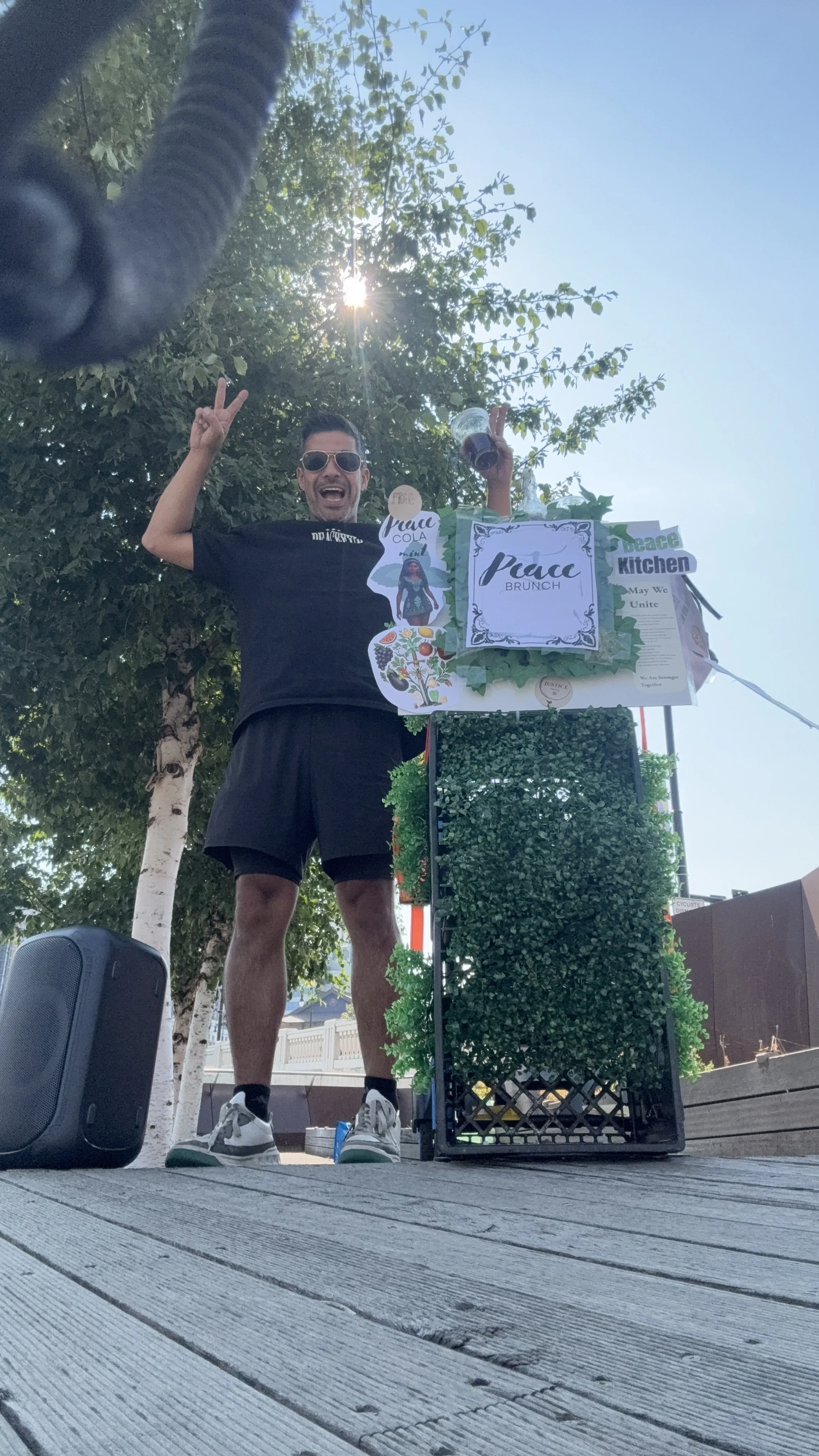A cheerful man in sunglasses and black shorts standing outdoors on a wooden deck, raising two fingers in a peace sign, next to a decorated stand with a sign that says 'Peace Brunch'. The stand has green foliage and various signs, with a tree and blue sky in the background.