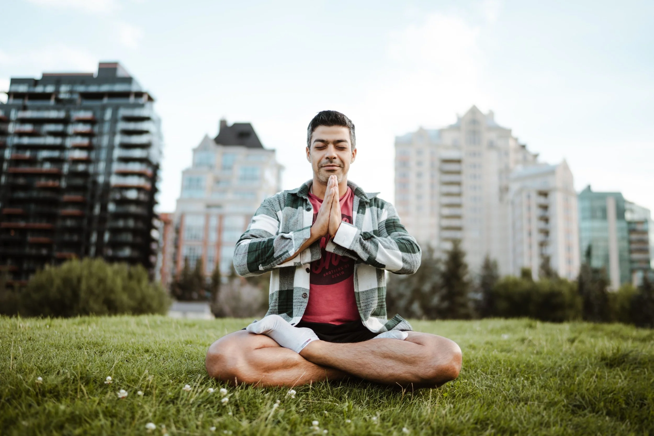 A man practicing meditation outdoors on a grassy area with city buildings in the background.