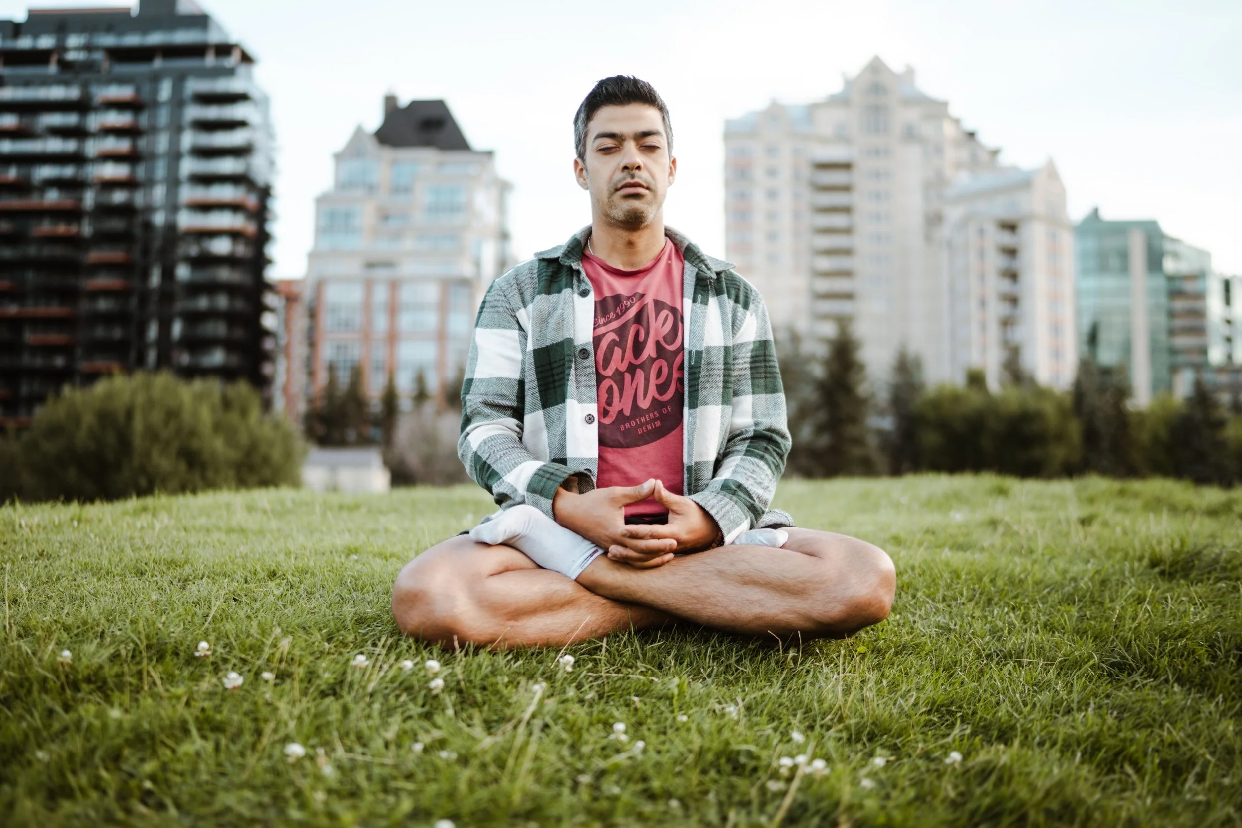 A man sitting cross-legged on grass in a meditative pose with eyes closed, wearing a red t-shirt and a plaid shirt, with a cityscape of tall buildings in the background.