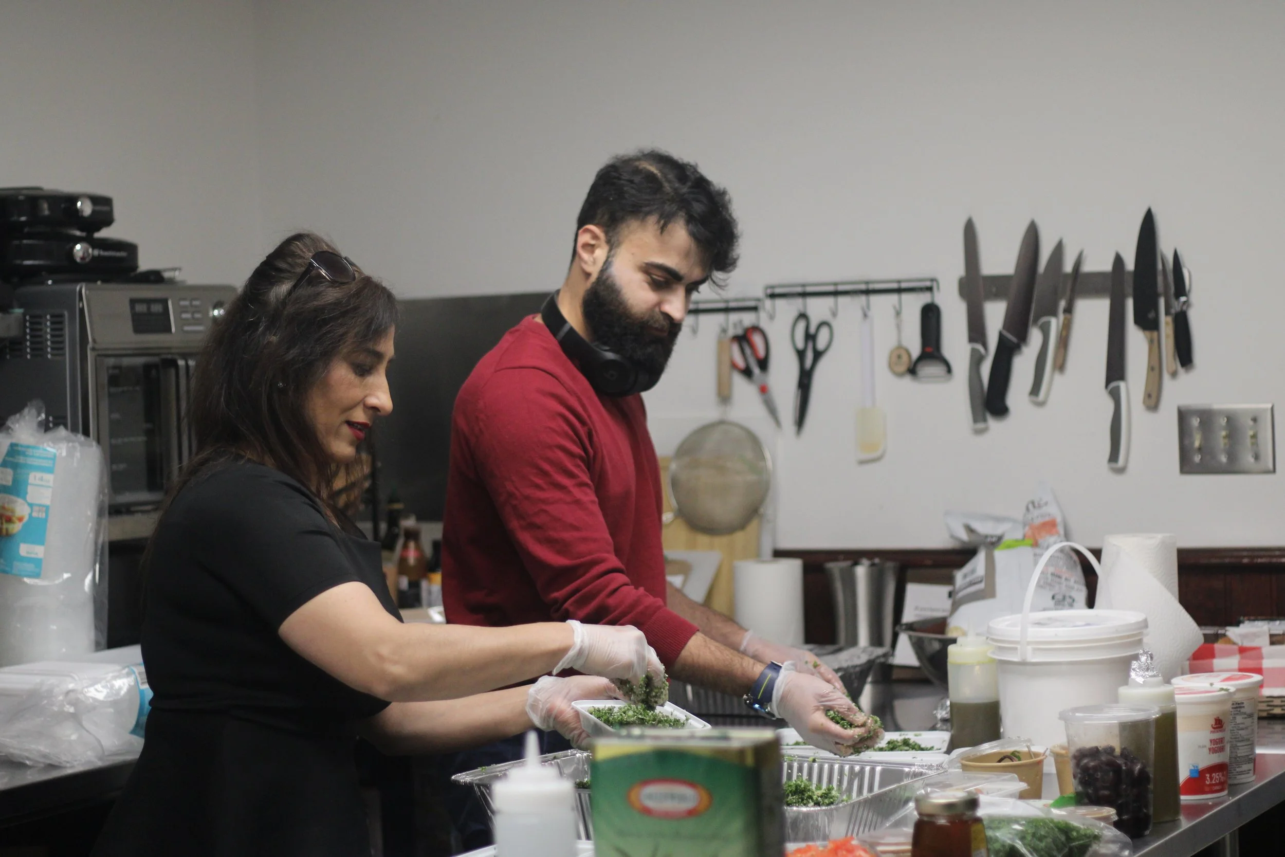 Two people preparing food in a kitchen, wearing gloves, with various kitchen utensils hanging on the wall.