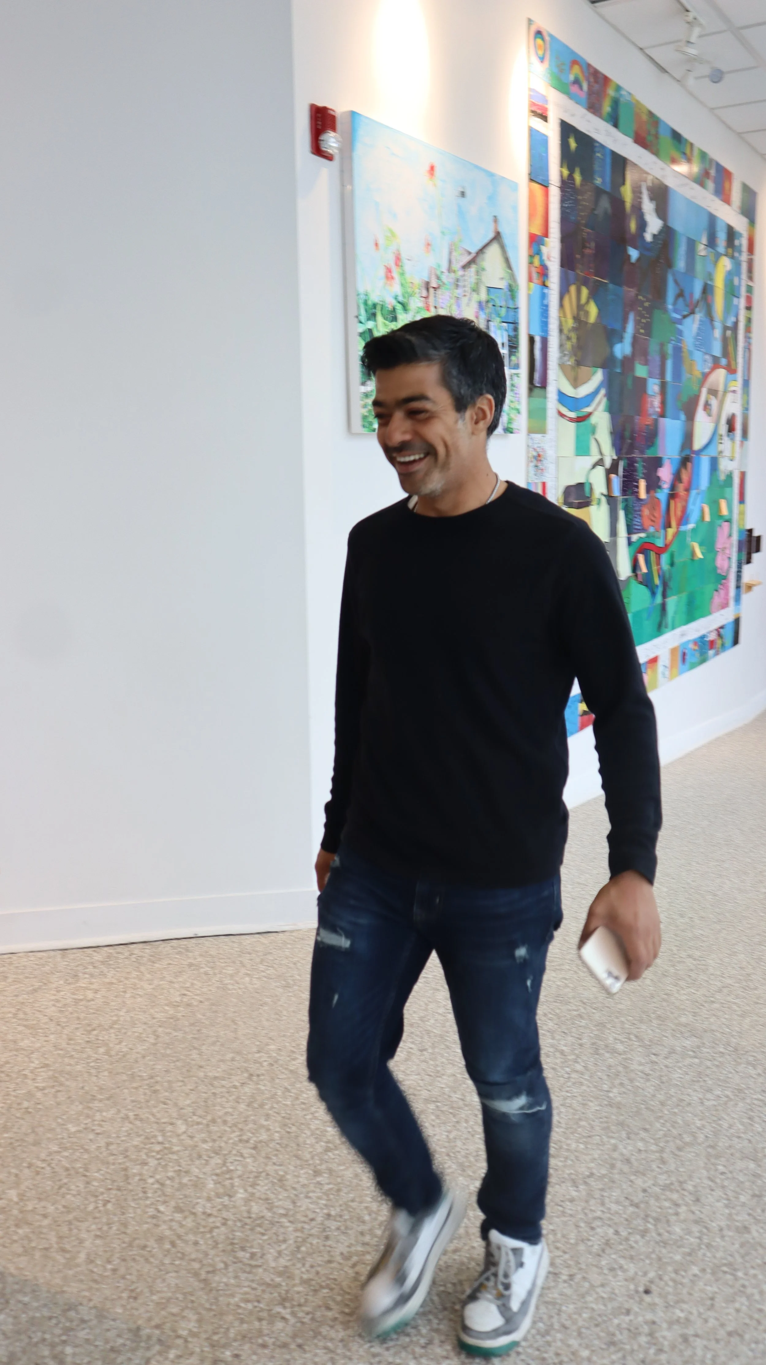 A man with dark hair, wearing a black long-sleeve shirt, ripped jeans, and white sneakers, walking and smiling in an art gallery with colorful artwork on the wall behind him.