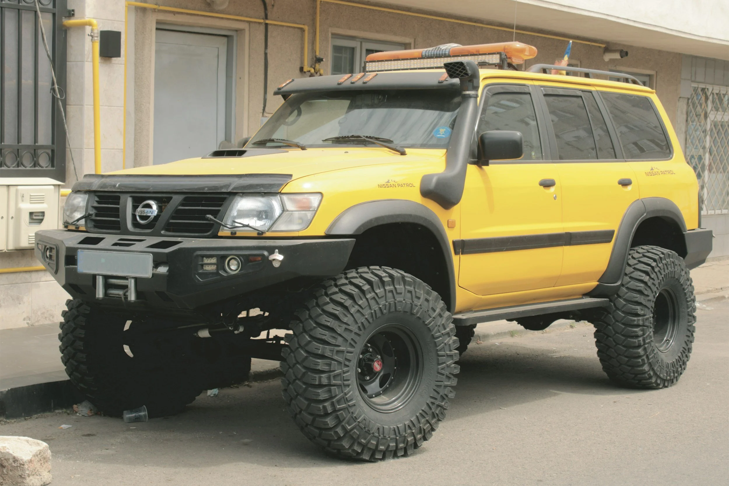 A yellow Nissan Patrol police vehicle with large off-road tires, black bumper, roof lights, and equipment, parked on a city street.