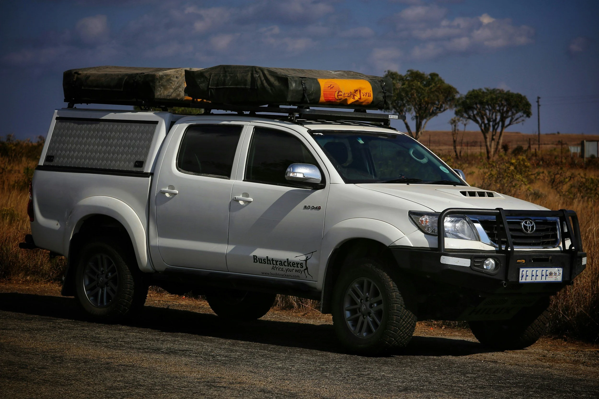White Toyota pickup truck with off-road bumpers and a canopy, parked on a dirt road in a dry landscape with sparse trees, carrying roof-mounted gear and a cargo bag.