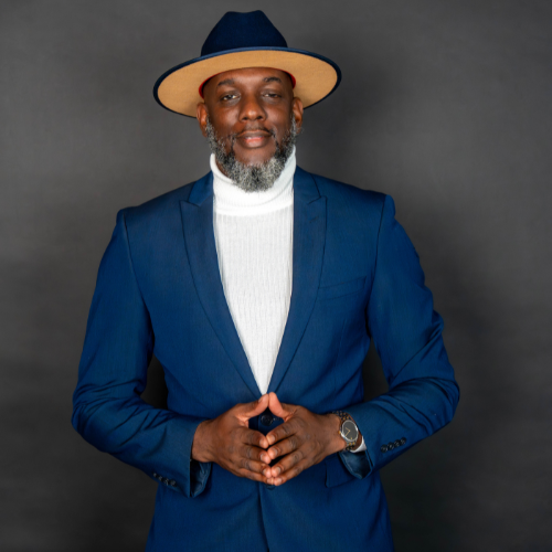 Man in a blue suit, white turtleneck, and wide-brimmed hat posing against gray background.