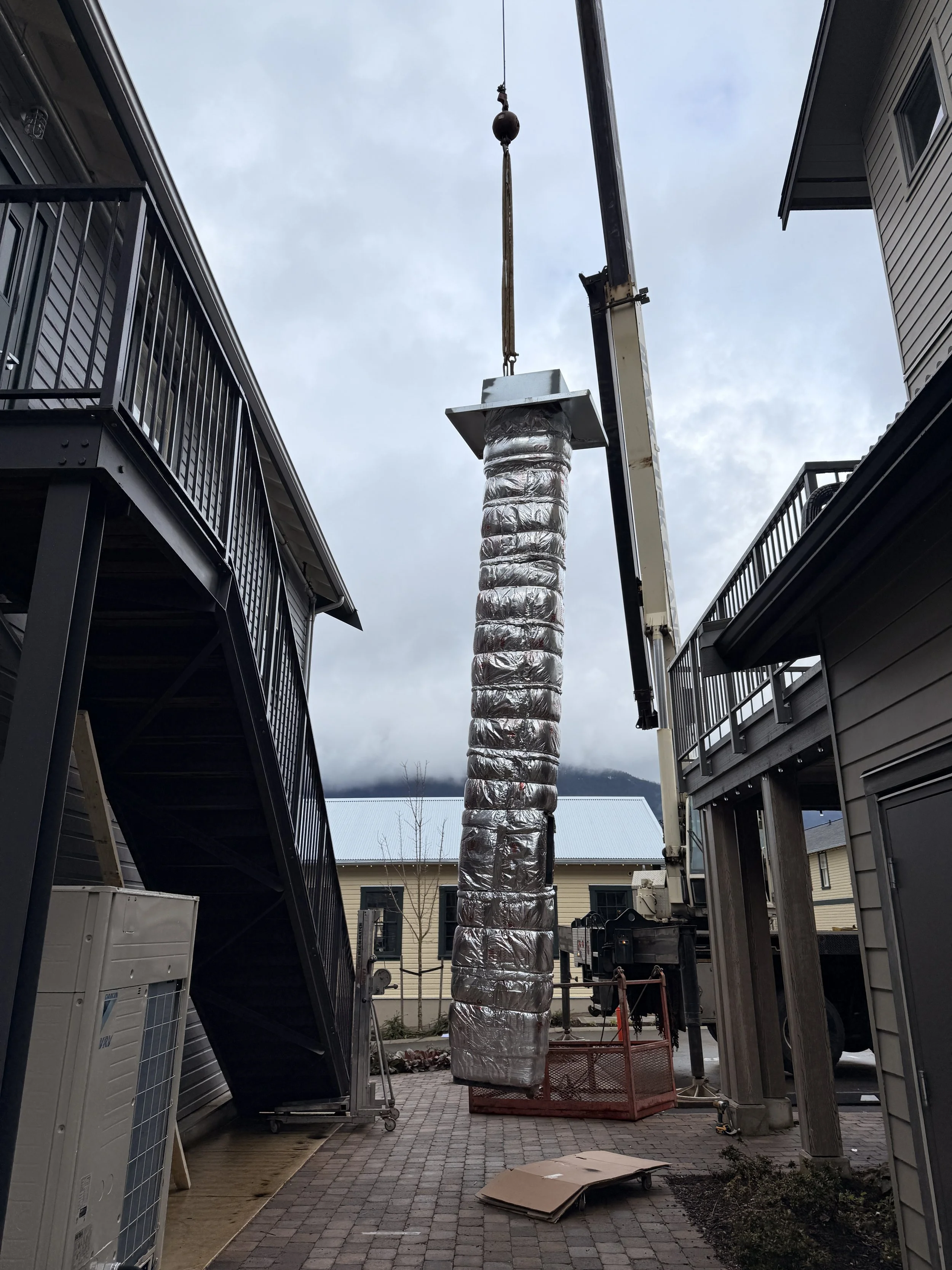 A tall duct wrapped in shiny silver insulation being installed between two residential buildings using a crane.