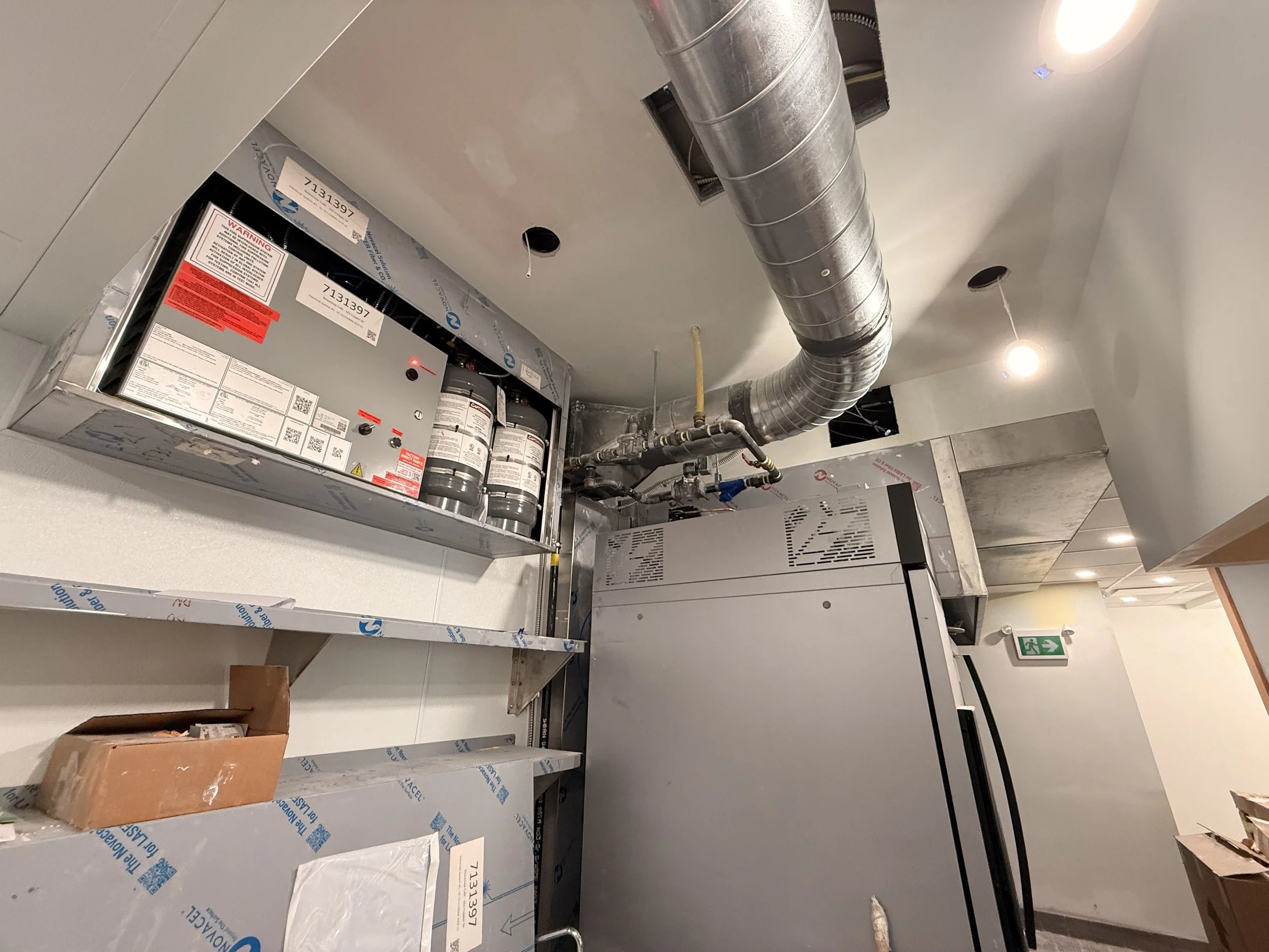 Mechanical room with ventilation ductwork, an electrical panel, fire suppression system, and a gray refrigerator.