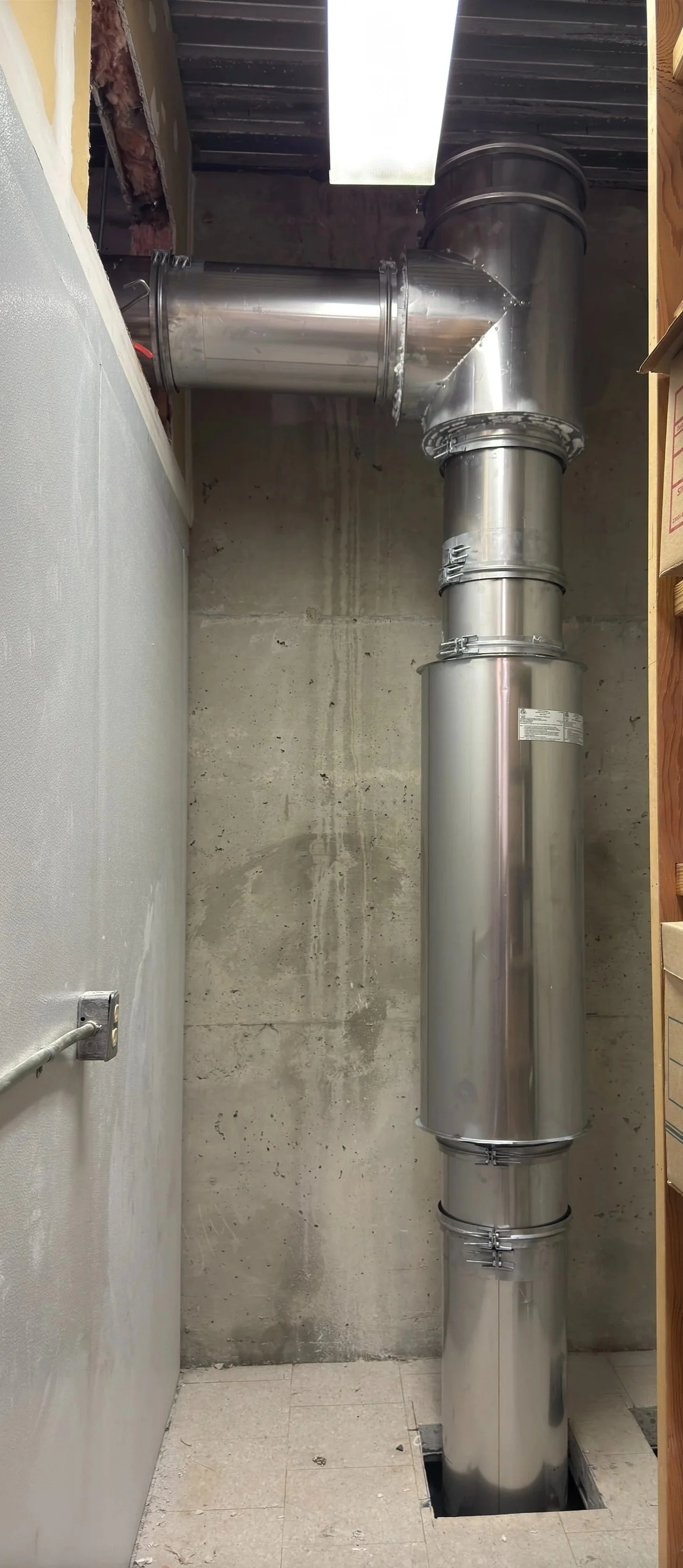 Marble floor with a rectangular hole around a large, shiny, gray metal vent pipe running vertically from the floor to the ceiling in a basement or utility room with concrete wall and ceiling, and a duct attached to the pipe.