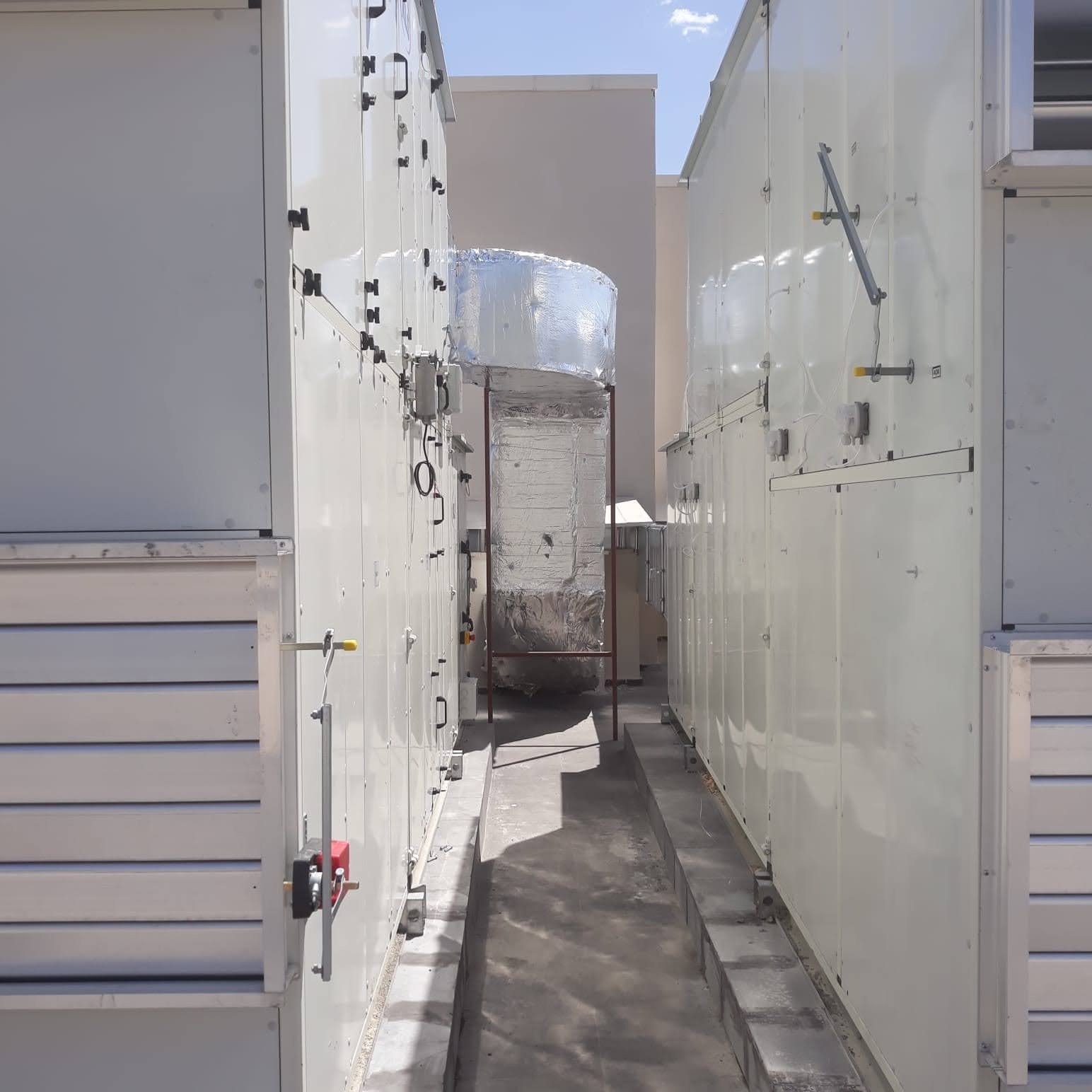 View of two large white HVAC units installed on a building's rooftop, with a duct connection wrapped in reflective insulation material between them, under a partly cloudy sky.