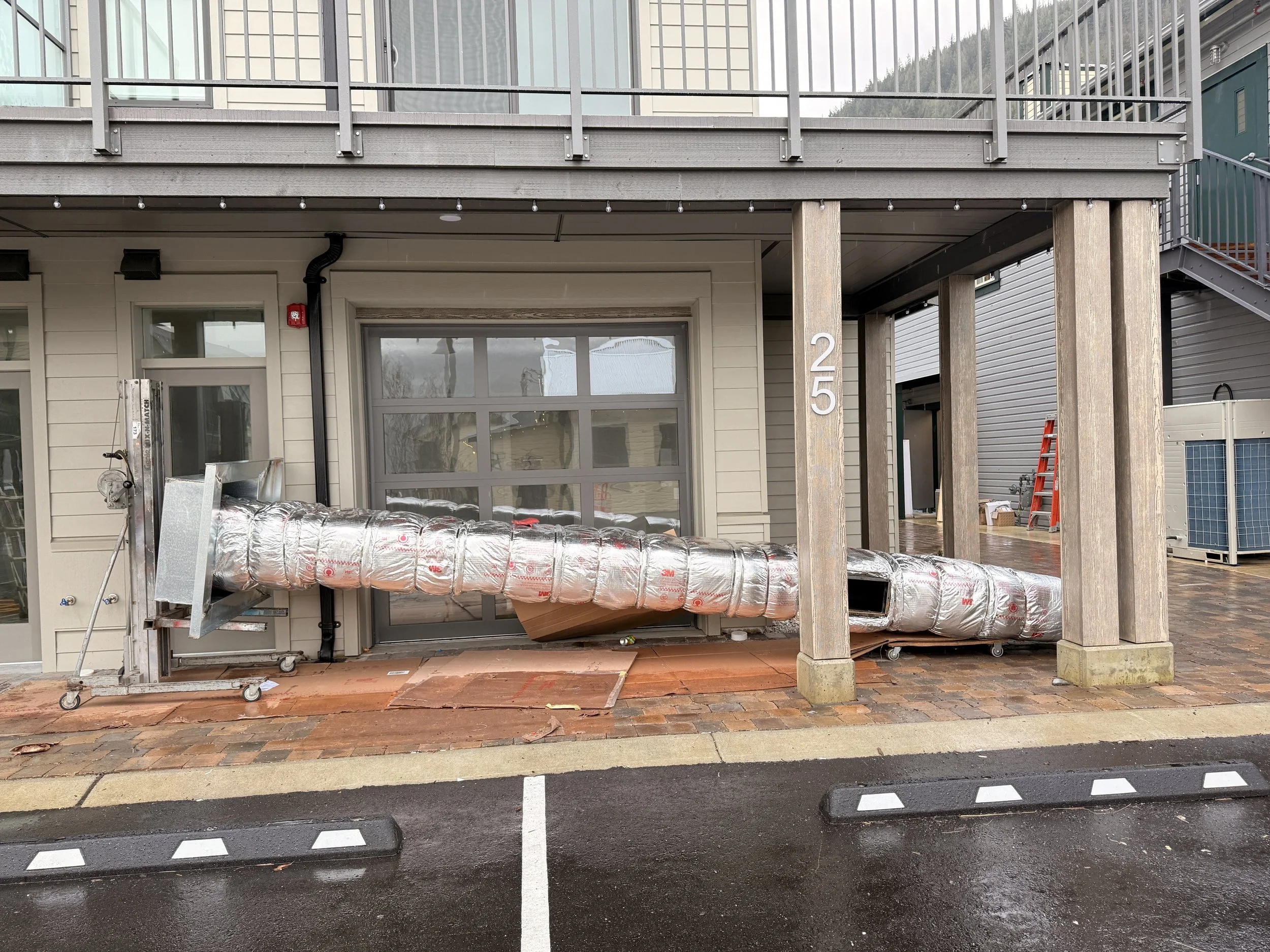 Construction site with HVAC ductwork connected to a building, with a parking lot in the foreground and the building number 25 on a wooden post.