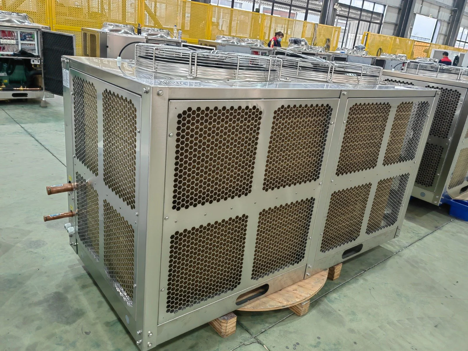 Large industrial air conditioning or refrigeration unit with metal honeycomb panels, copper pipes, and wire guards in a factory or warehouse setting.