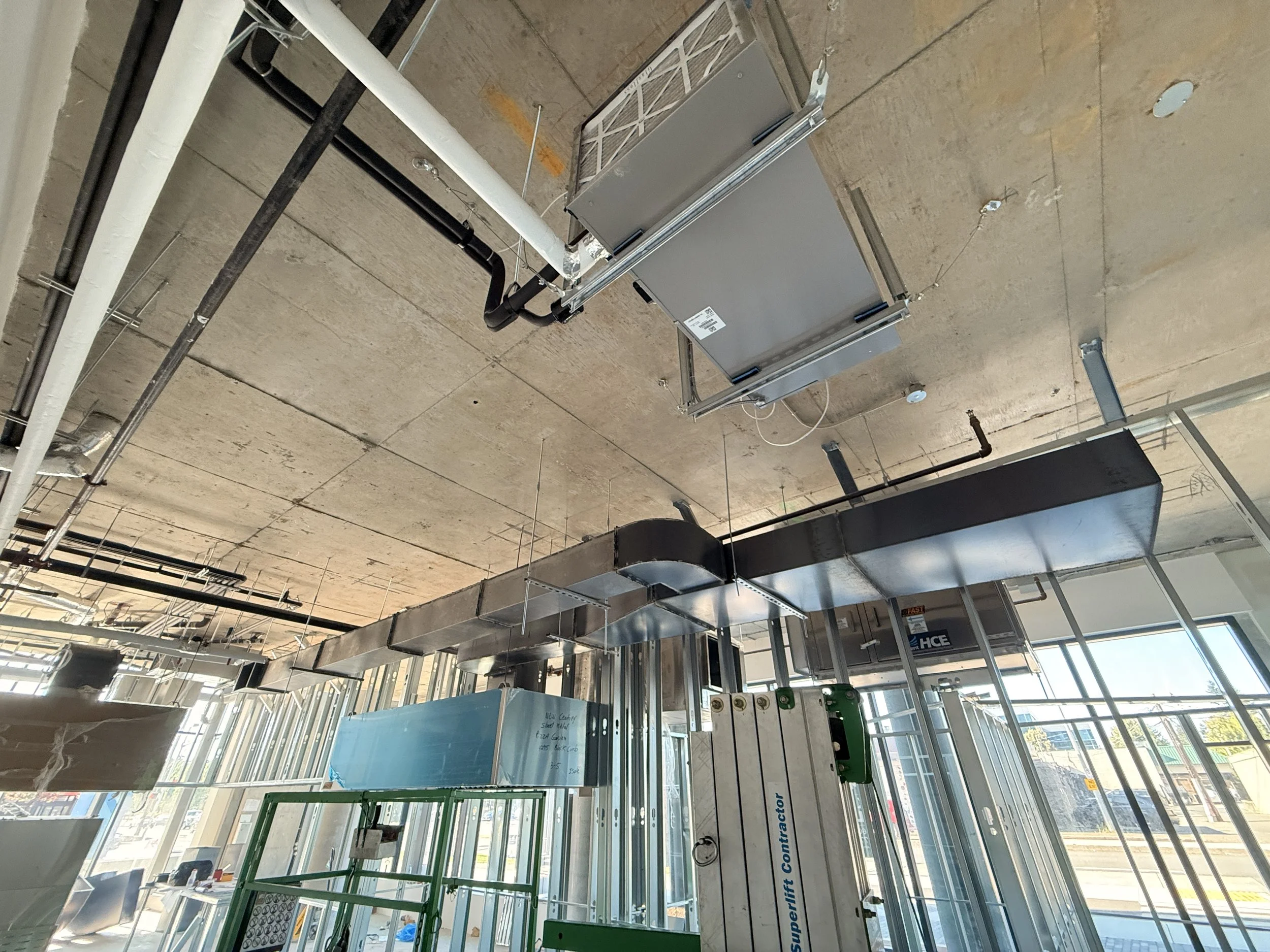 Construction site interior with exposed ceiling, ductwork, pipes, and metal framing for walls or equipment.