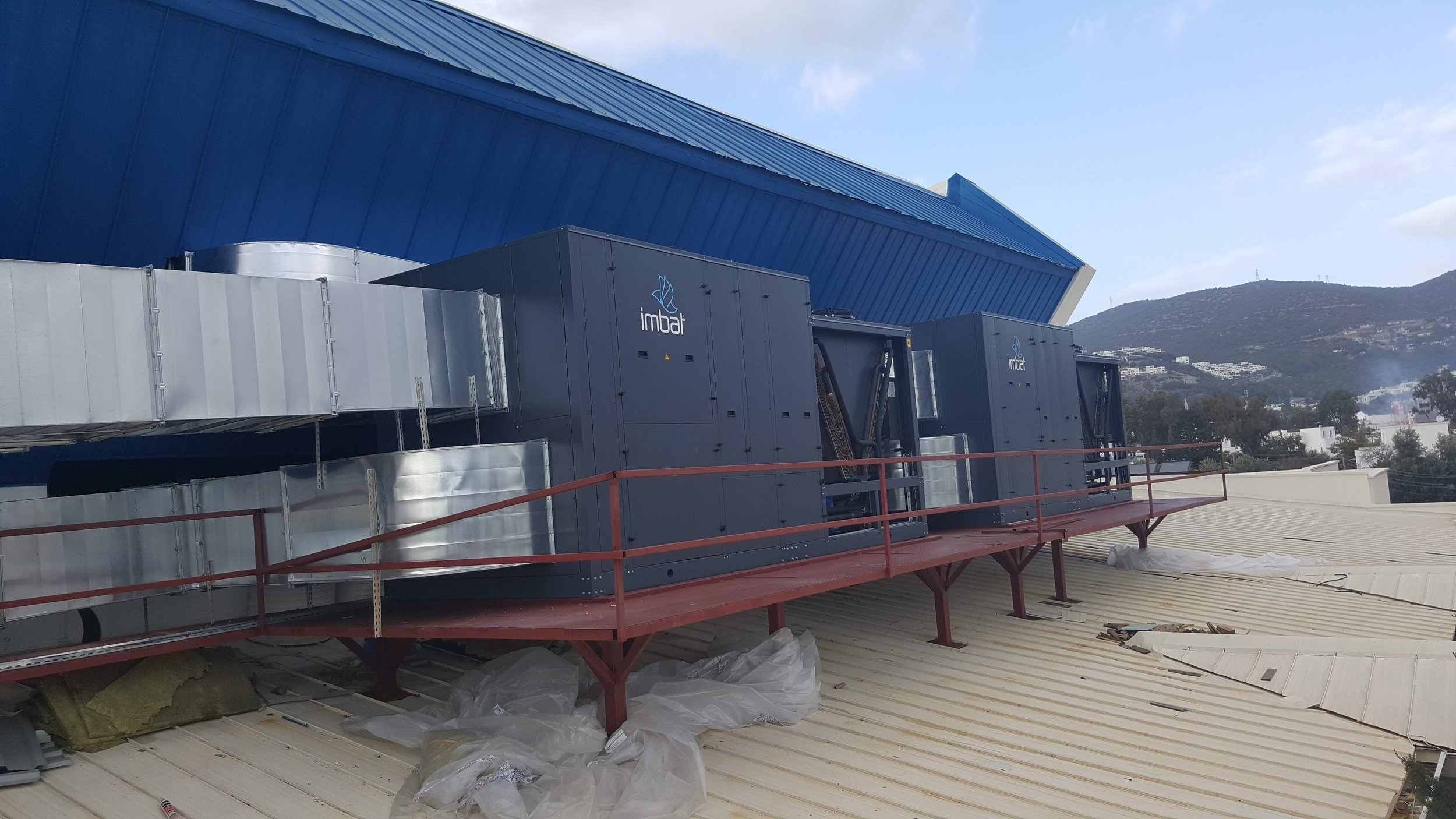 Commercial rooftop with HVAC units labeled 'imbat' on a metal balcony, overlooking a hilly landscape with a blue and white building nearby.