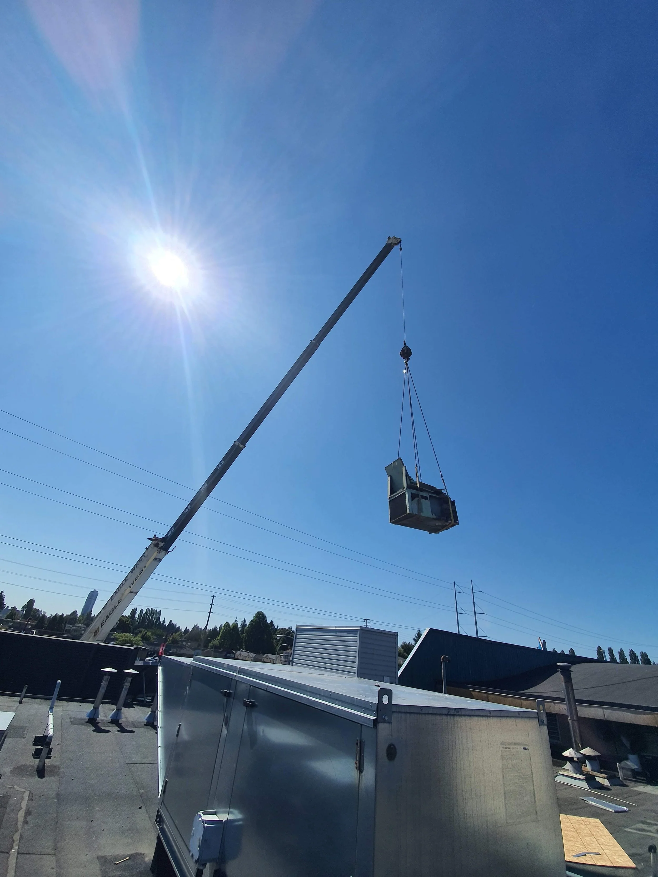 A crane lifting a large air conditioning unit or similar equipment in an outdoor industrial area under a clear blue sky with the sun shining brightly.