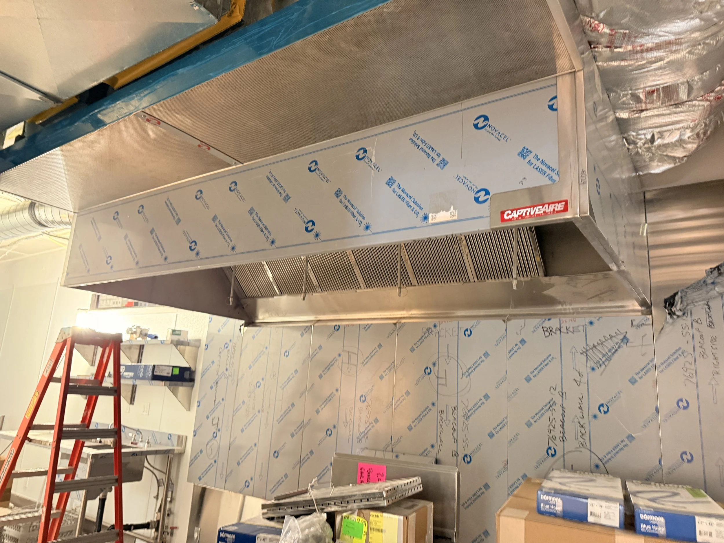 Commercial kitchen with a stainless steel range hood, a red ladder, and boxes on a counter, walls covered in protective blue film.