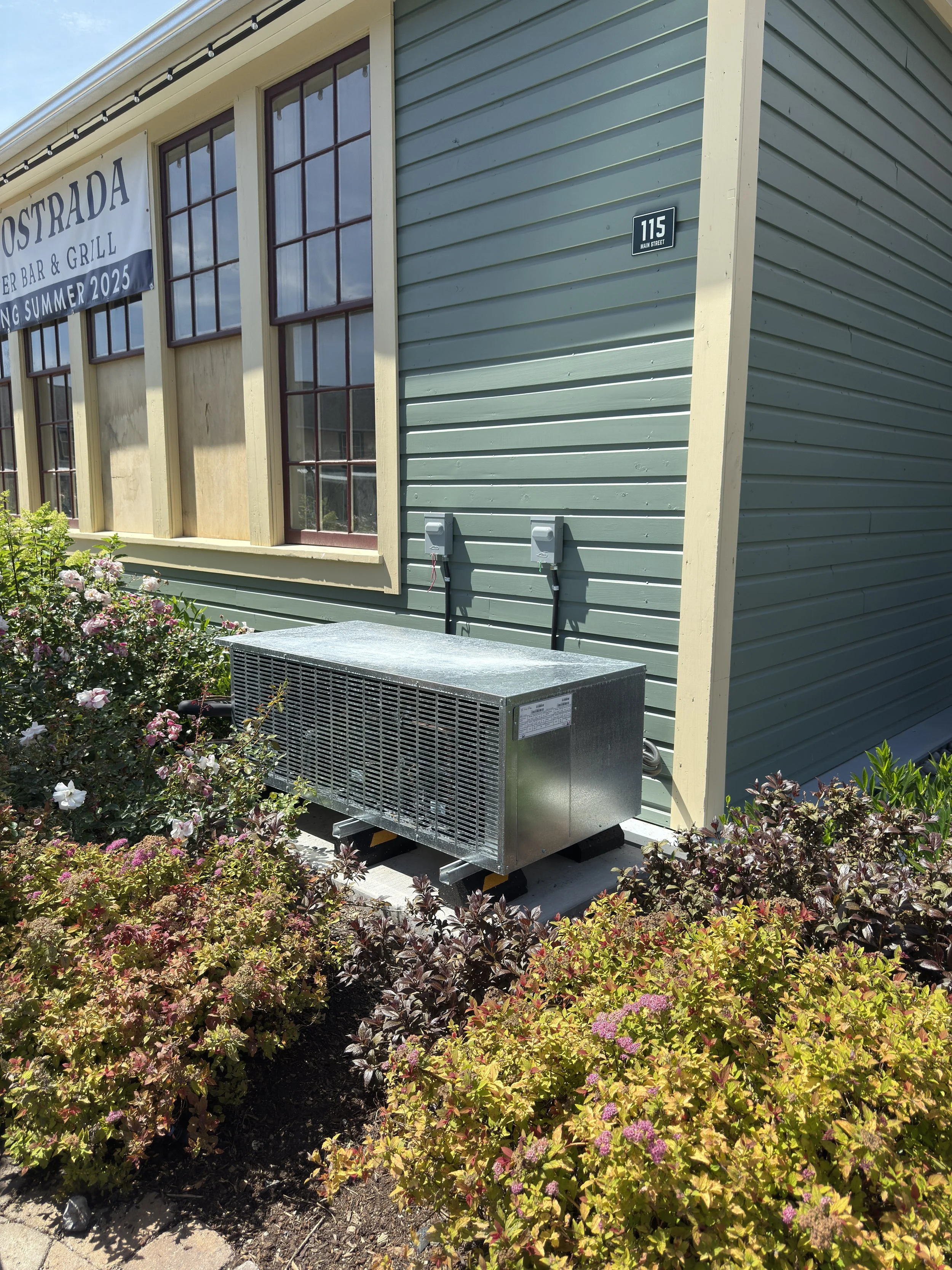 Exterior of a building with green siding, large windows, and a sign that reads "Ostrada Sports Bar & Grill." There are two electrical boxes on the wall, and a metal cooling unit placed on the ground near flowering bushes.