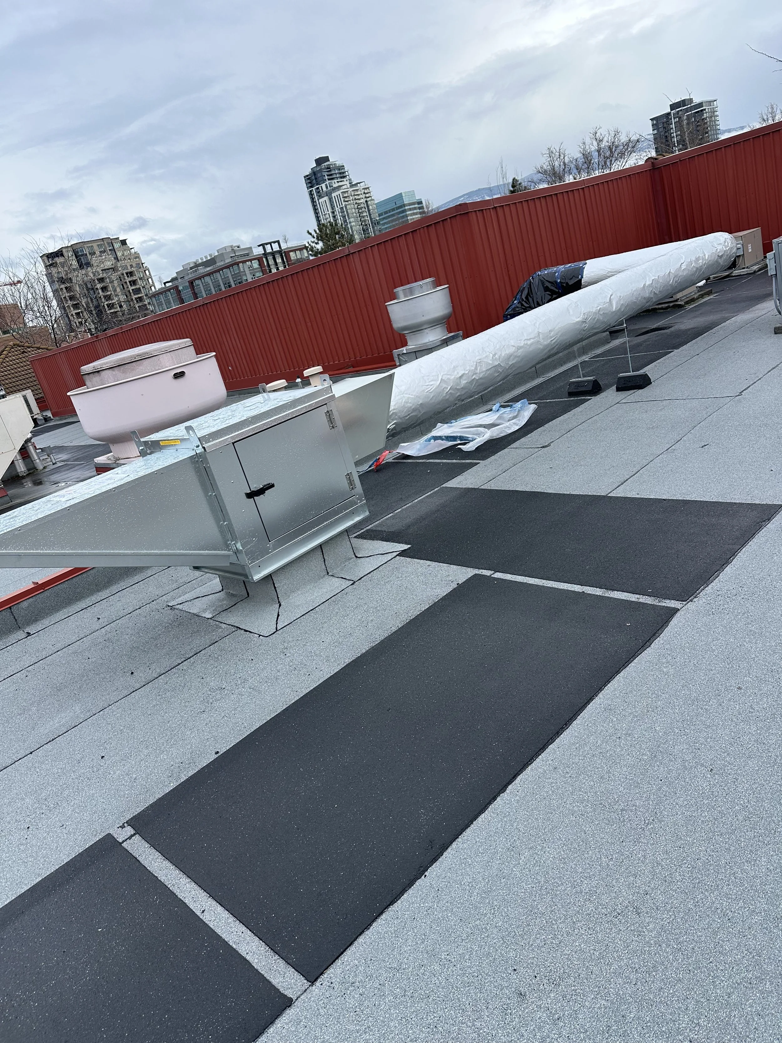 Rooftop with HVAC system, ducts, vents, and black rubber security mats, with city buildings and cloudy sky in background.