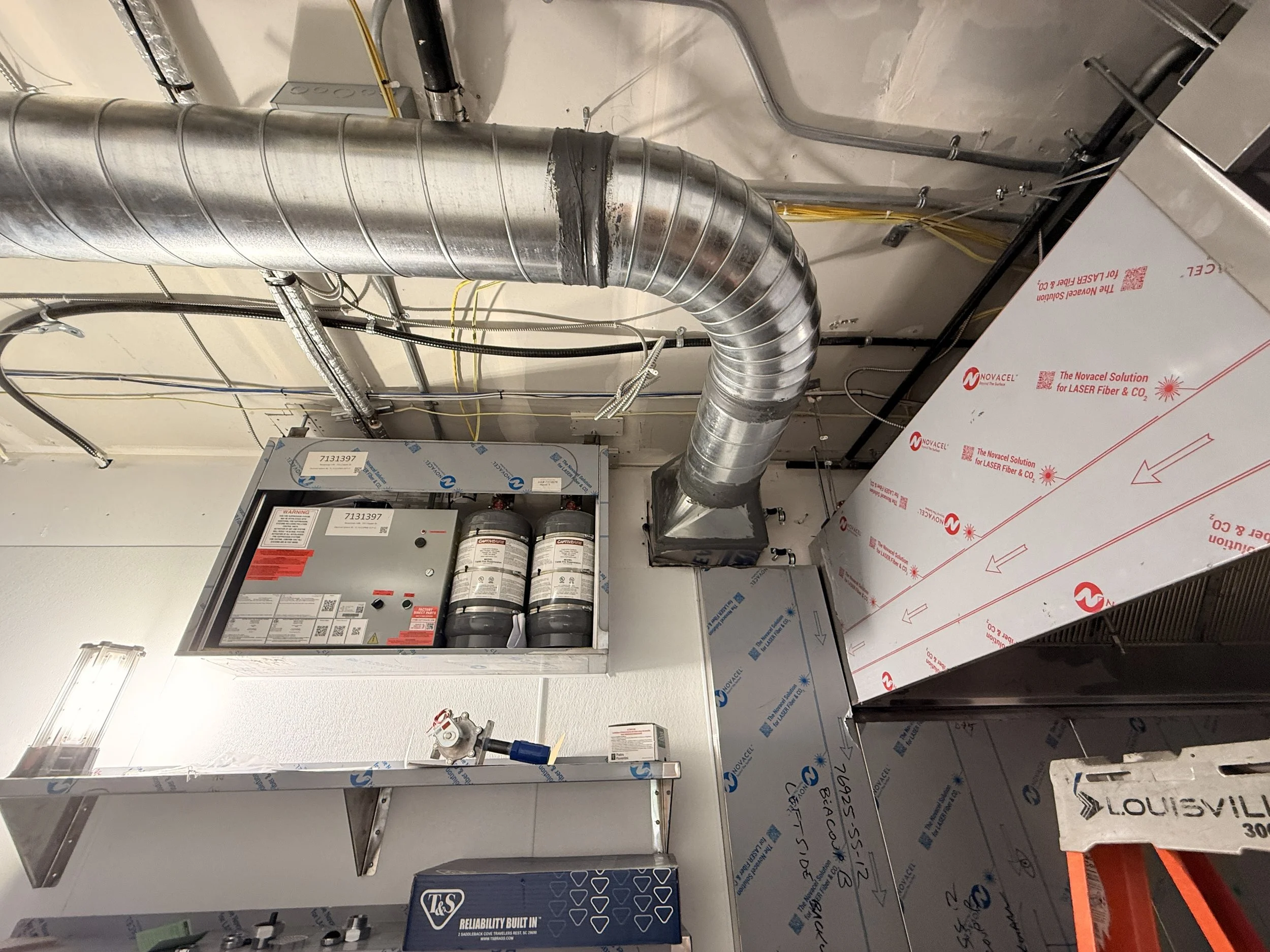 Ceiling-mounted HVAC system with large silver ductwork, a fire safety panel with two fire extinguishers, and various wiring and pipes in a commercial or industrial setting.
