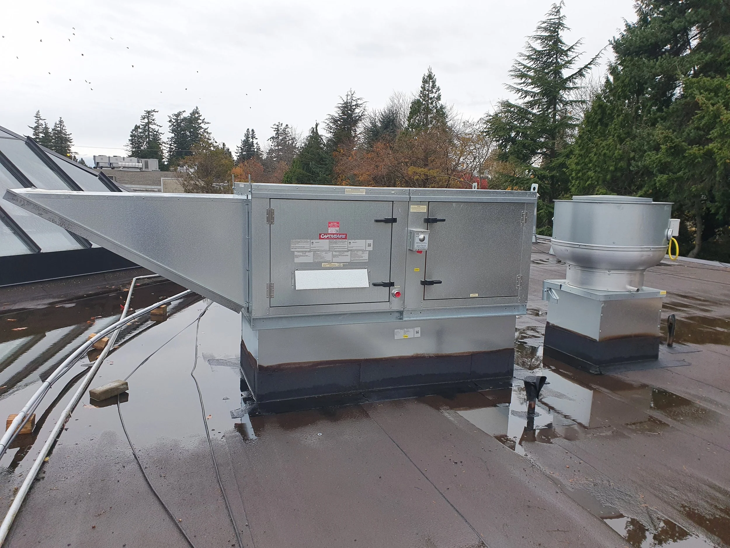HVAC rooftop unit on a flat roof with trees and cloudy sky in the background, and some water puddles on the roof surface.
