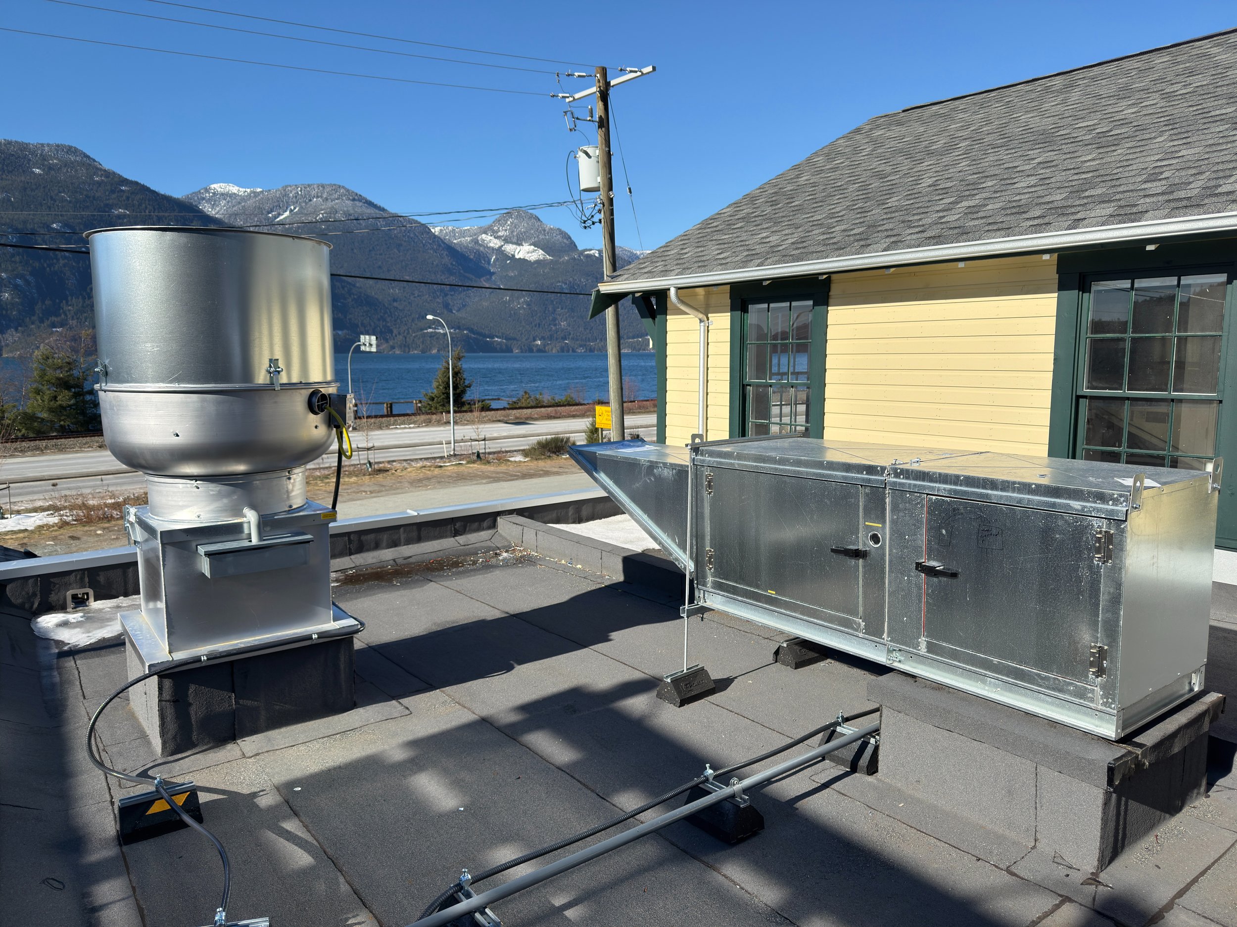 View of a rooftop with a large metal HVAC unit and a yellow house with green window frames, in front of a mountain and lake background, under a clear blue sky.