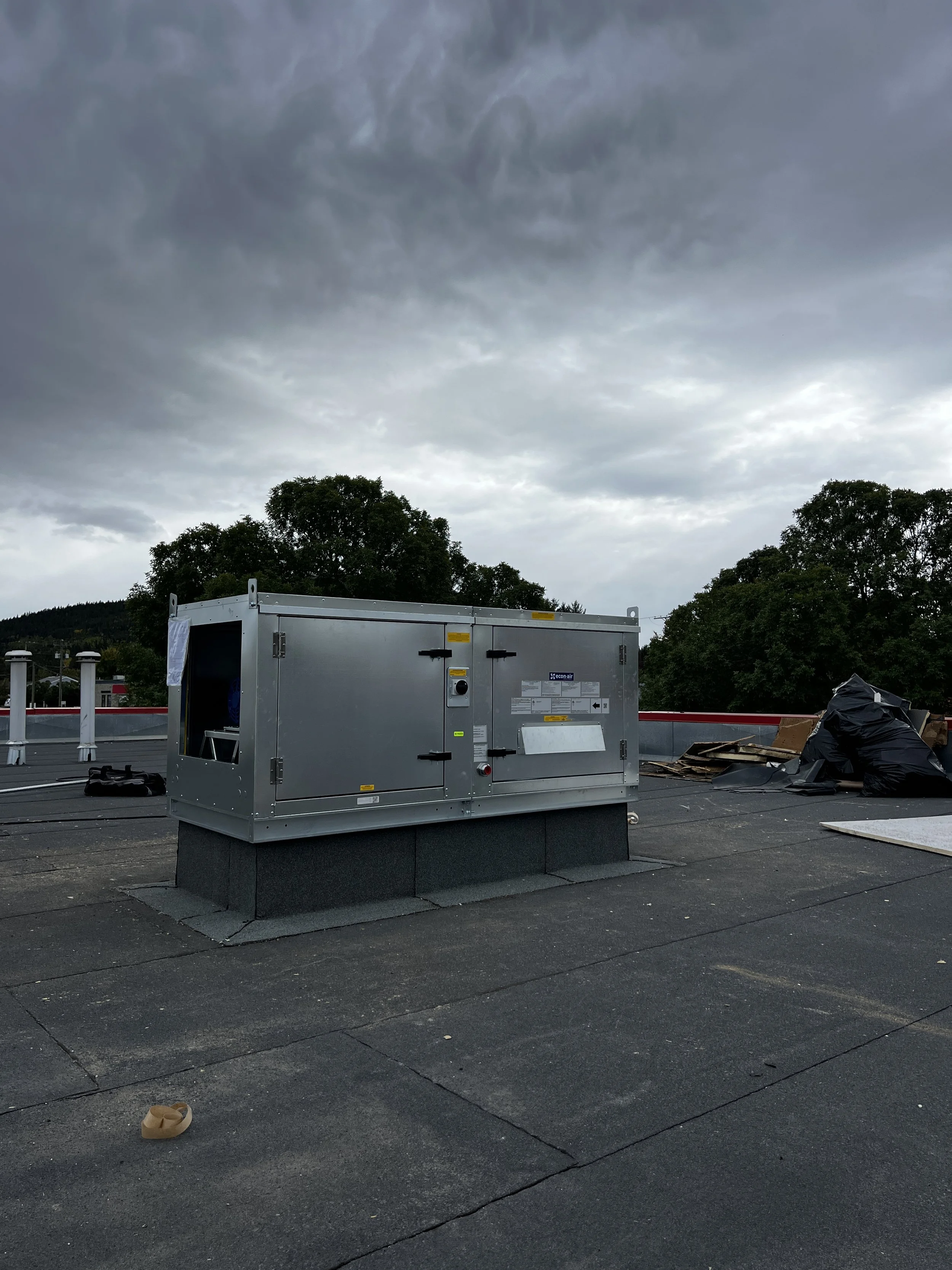 A rooftop scene featuring a large metallic HVAC unit with a cloudy sky overhead and trees in the background. There are some construction materials and black bags nearby.