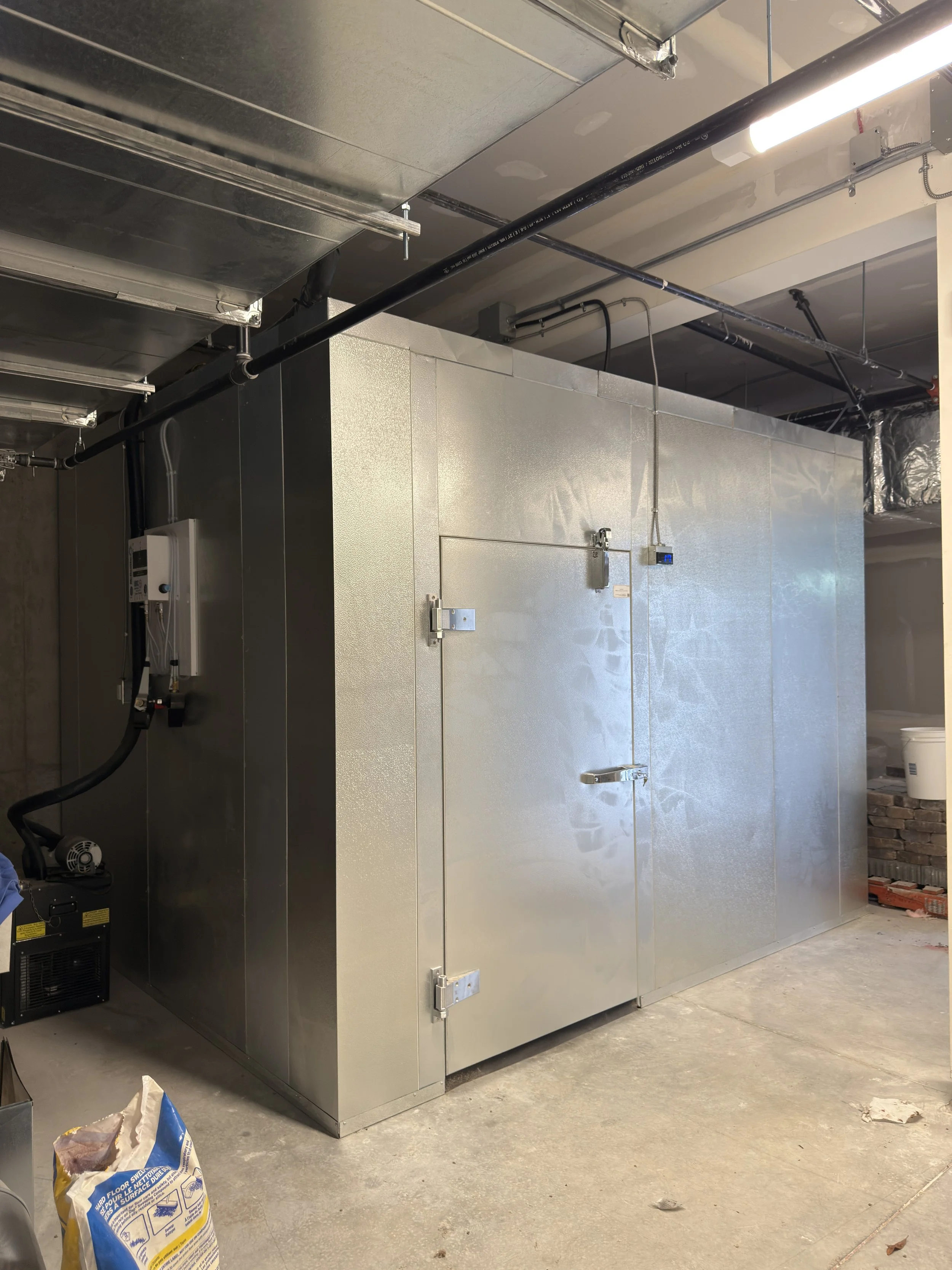 Large metallic industrial or commercial refrigeration unit or cold storage room in a garage or basement, surrounded by pipes and electrical wiring.