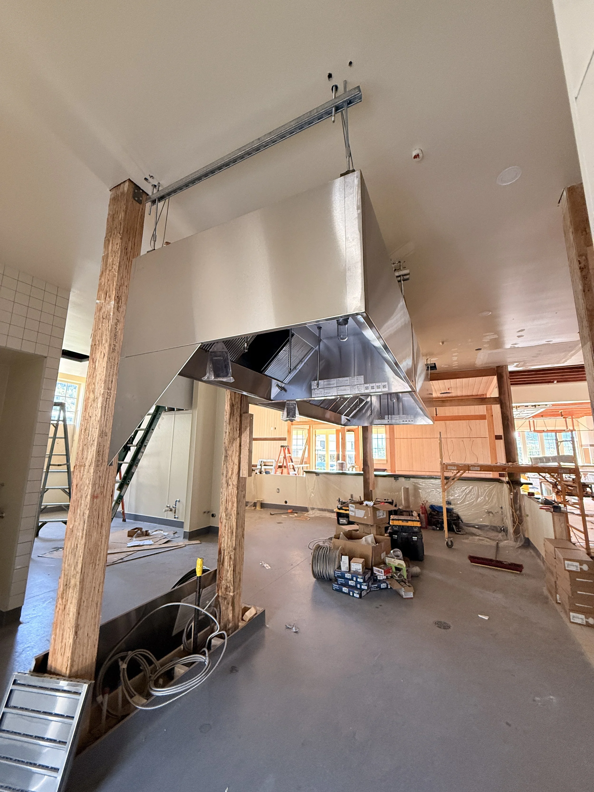 Construction site with unfinished interior, wooden support beams, a large suspended stainless steel kitchen hood, and construction tools and materials scattered on the floor.
