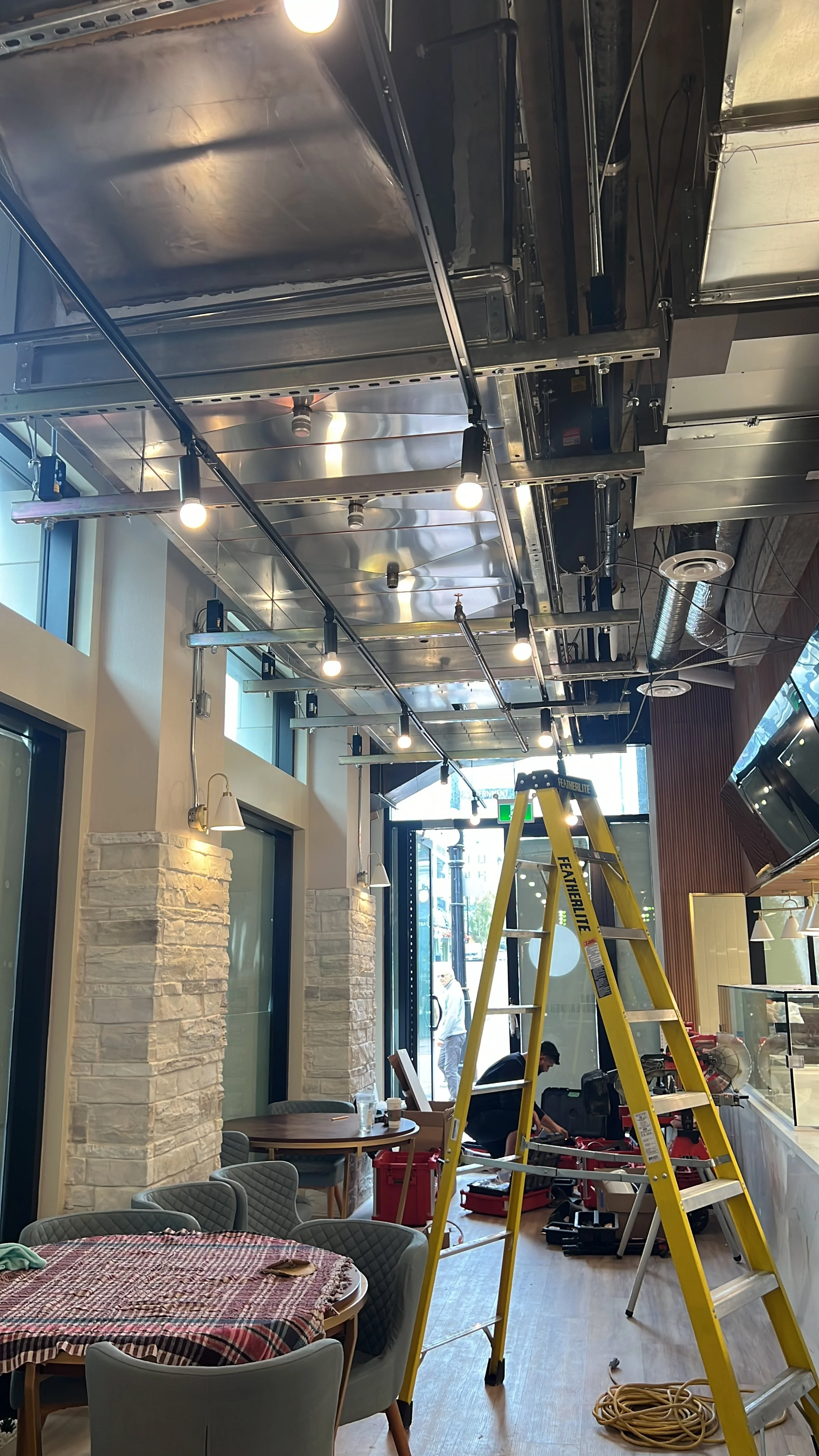 The ceiling of a restaurant under renovation with visible metal ductwork, track lighting with exposed bulbs, and a yellow ladder. There's a worker in the background working on equipment.