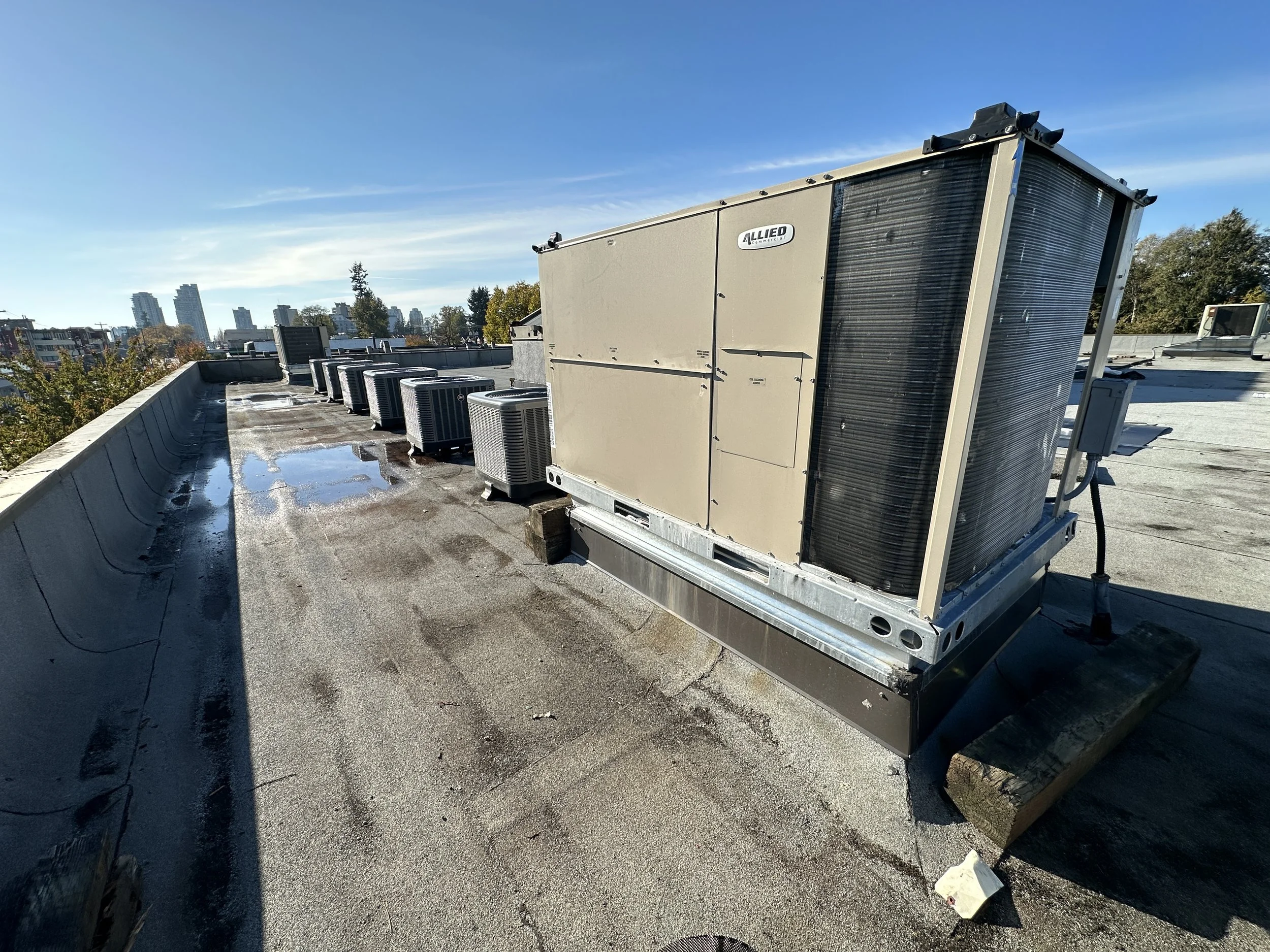 Rooftop with a large HVAC unit and multiple smaller air conditioning units, city skyline in the background, bright blue sky.