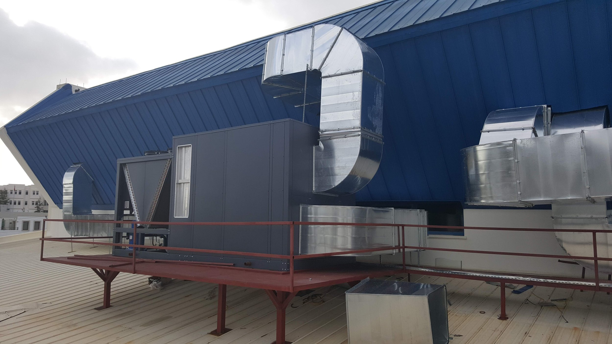 Rooftop HVAC system with metallic ducts and ventilation units installed on a beige metal roof with a blue house siding in the background.
