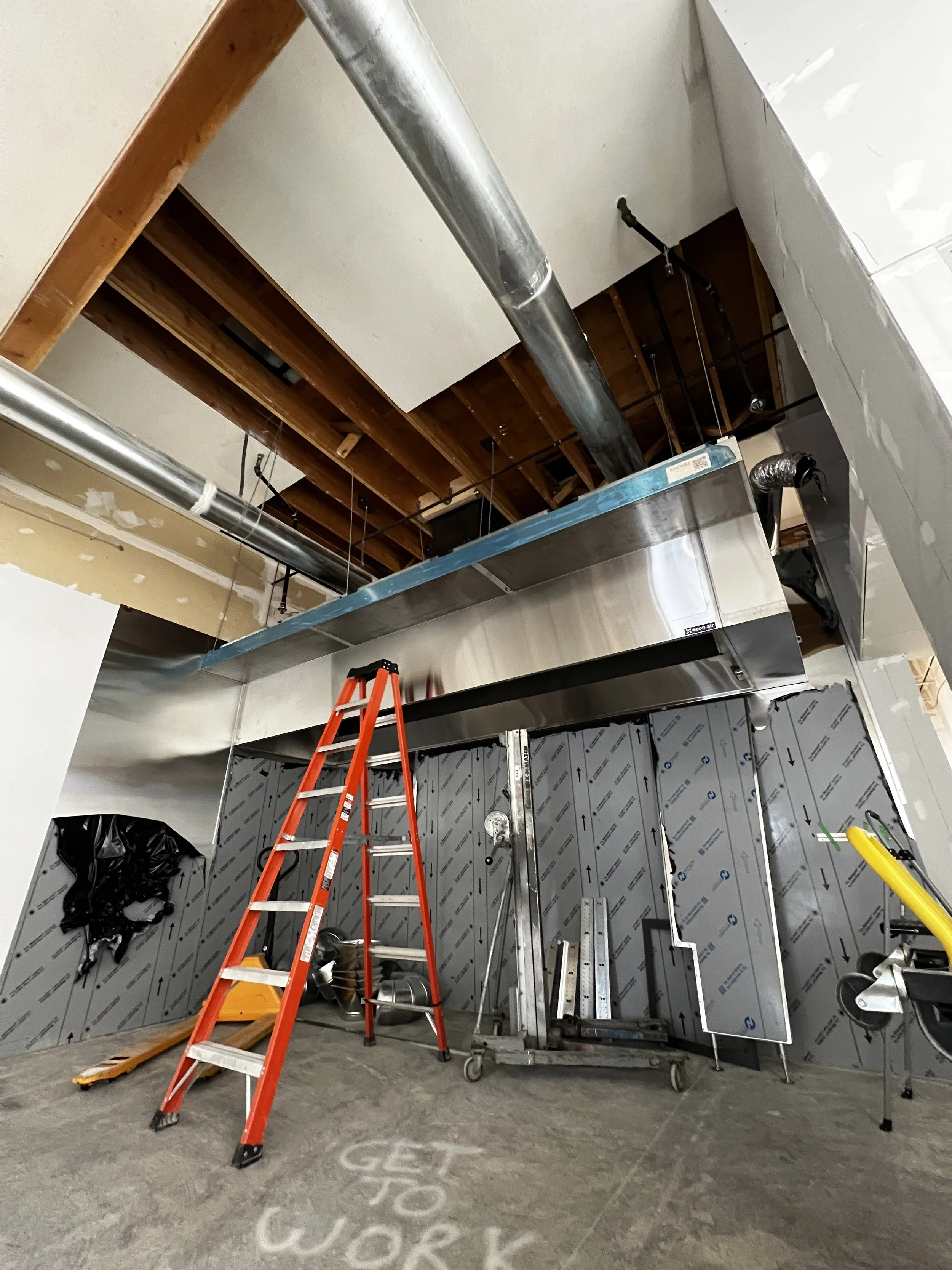 Construction site showing exposed ceiling with air ducts, a red ladder, construction tools, and partially installed kitchen cabinetry.