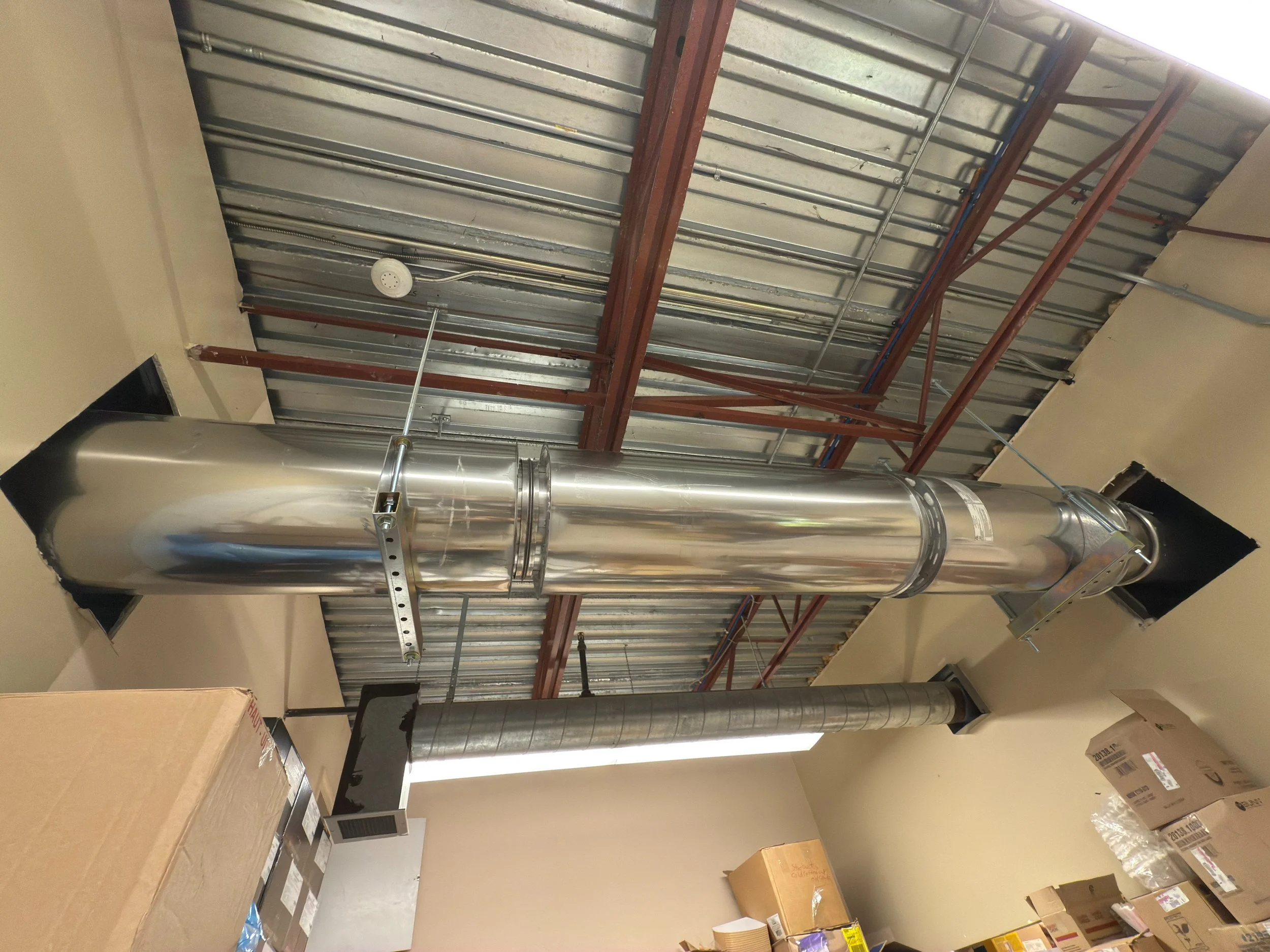 A large metallic industrial ventilation pipe installed in a utility or storage room with packages and boxes stacked nearby. The pipe runs vertically between the ceiling and the floor, with a smaller duct attached to the bottom.