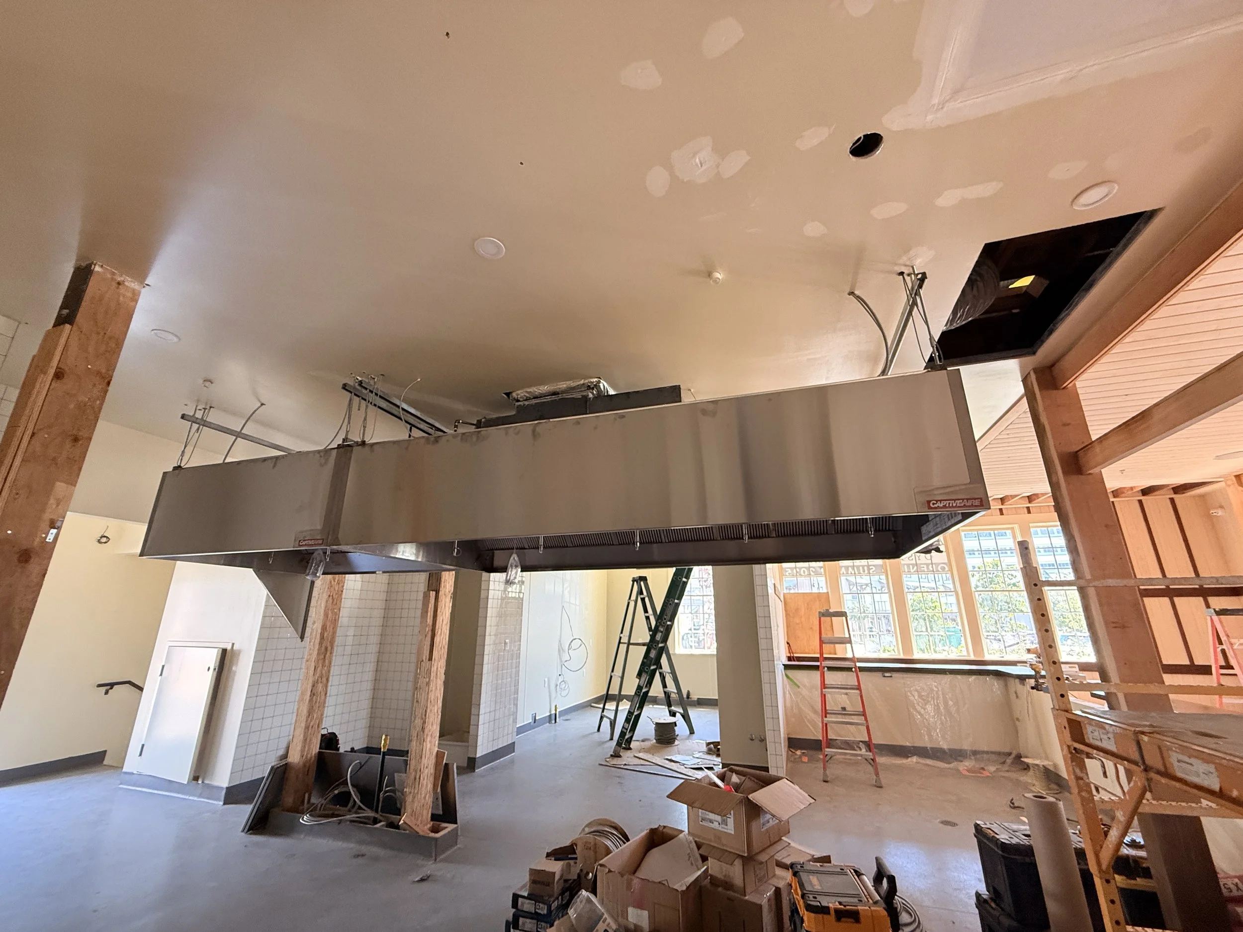 Construction site of a commercial kitchen with a large stainless steel ventilation hood, wooden support beams, ladders, and various tools and boxes on the floor.