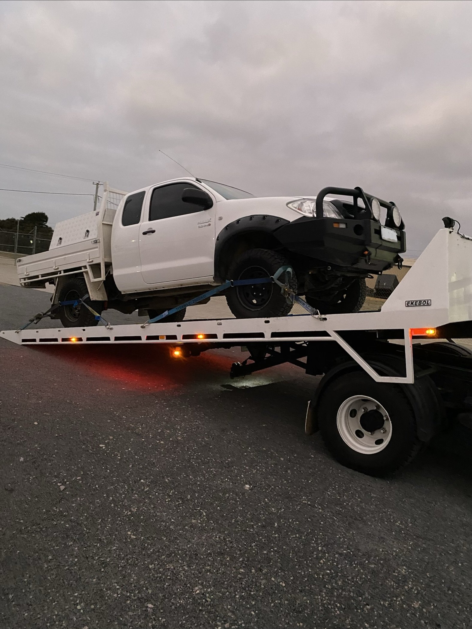 A white pickup truck loaded on a flatbed tow truck, with straps securing it, under a cloudy sky.