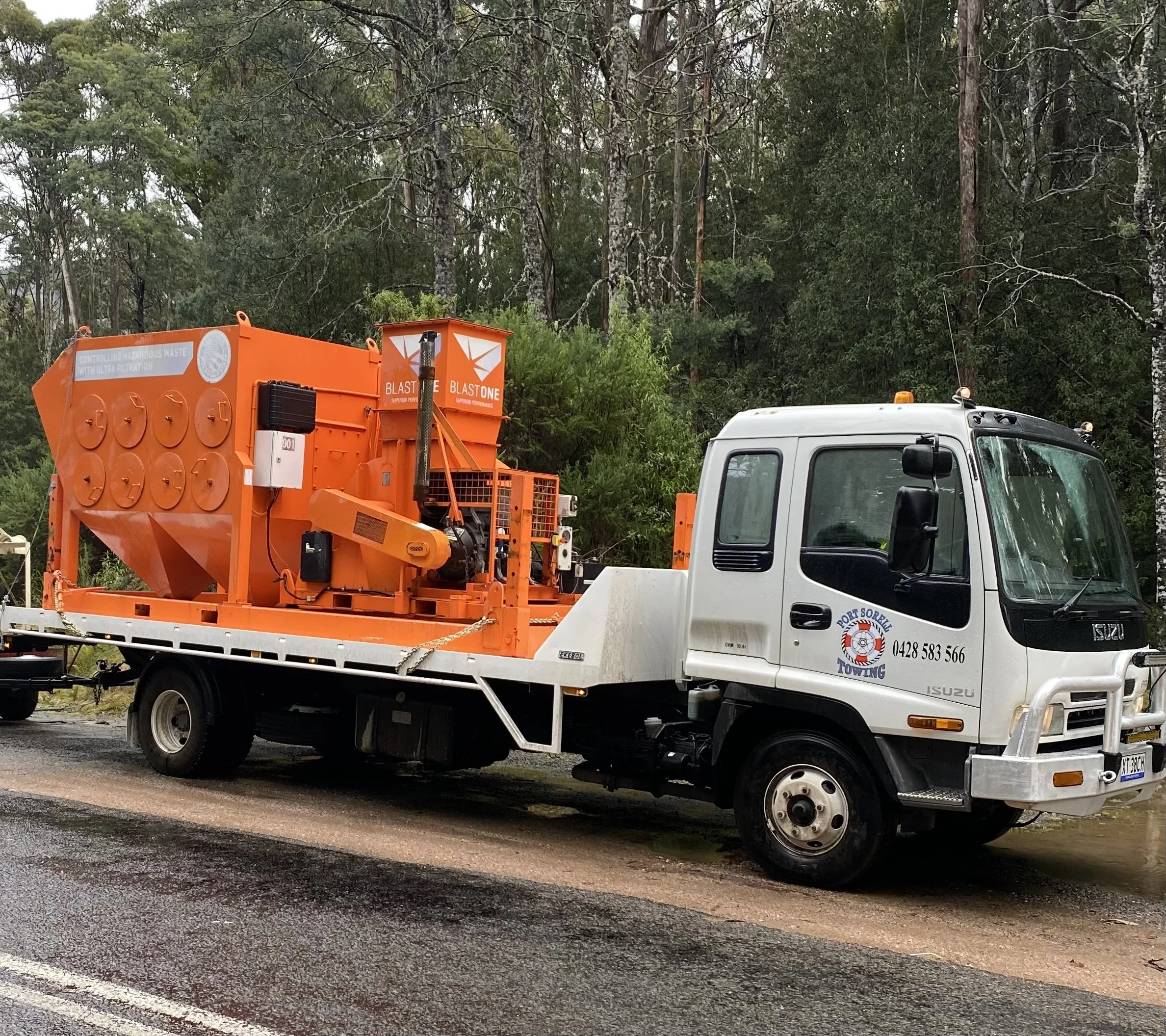 A white tow truck on a wet road, carrying an orange hazardous waste disposal machine, with a wooded area in the background.