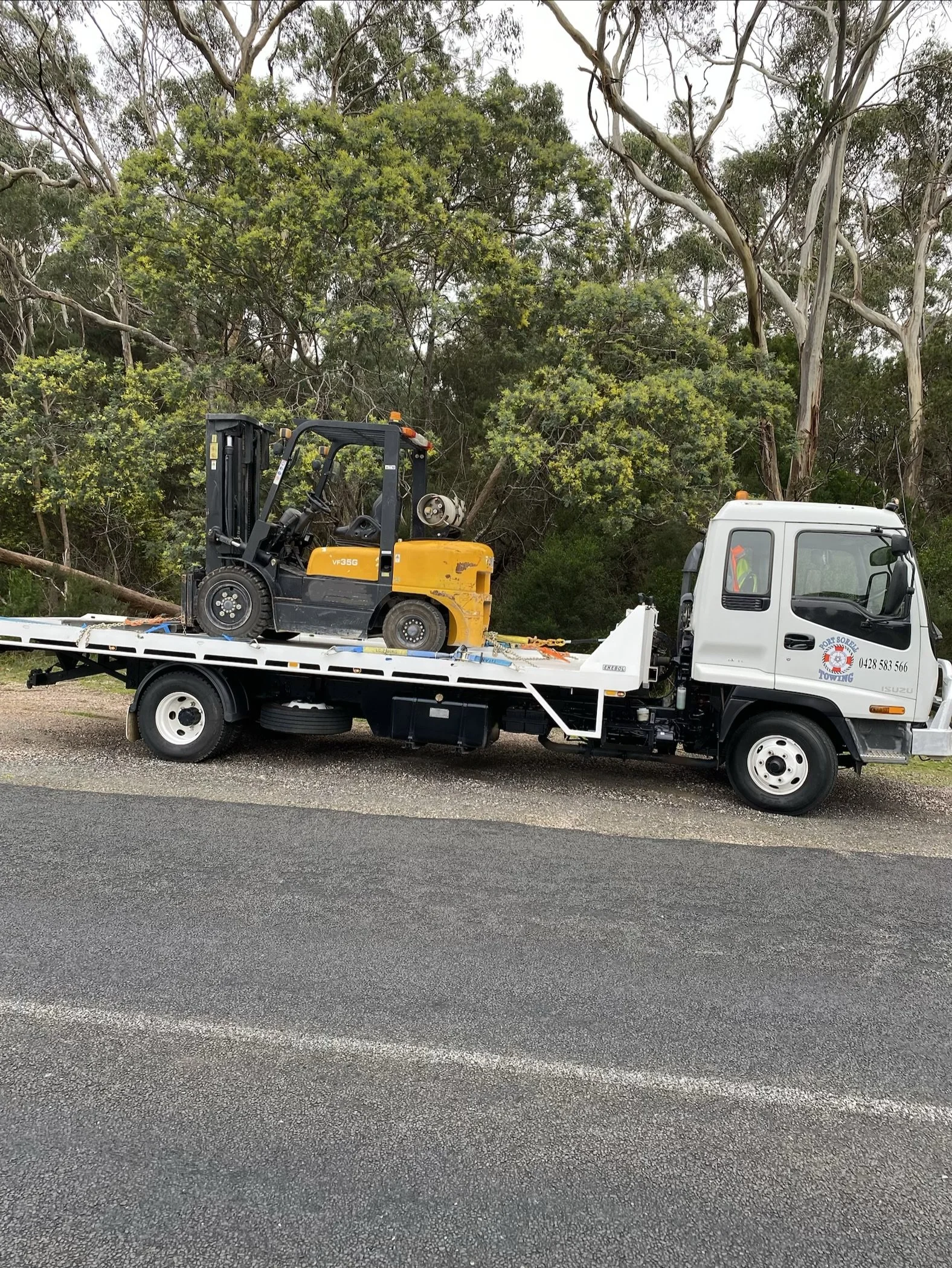 A white tow truck with a yellow forklift on its flatbed parked on the side of a road, with trees and green foliage in the background.