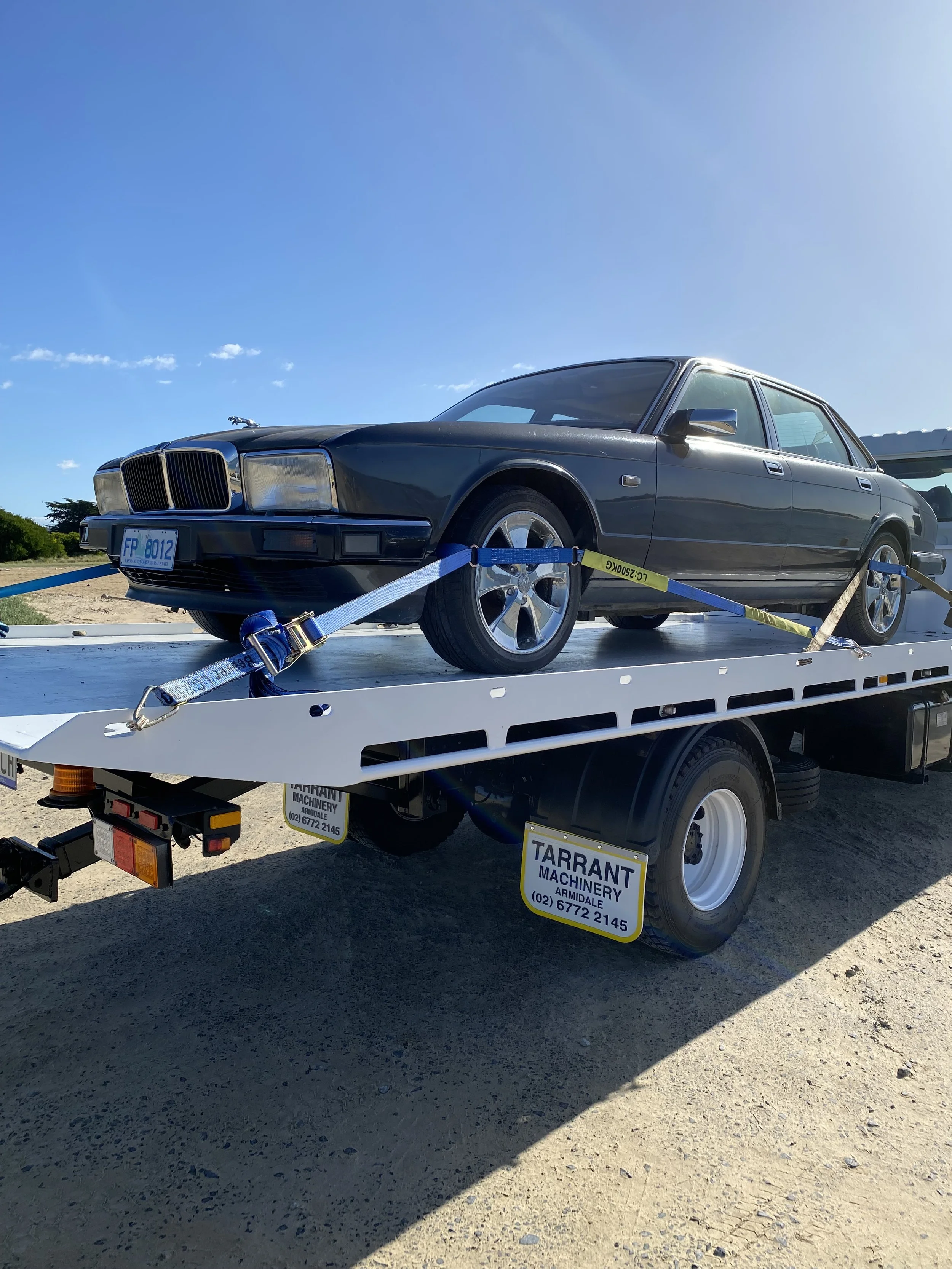 A vintage black sedan car on a flatbed tow truck, secured with blue straps, under a clear blue sky.