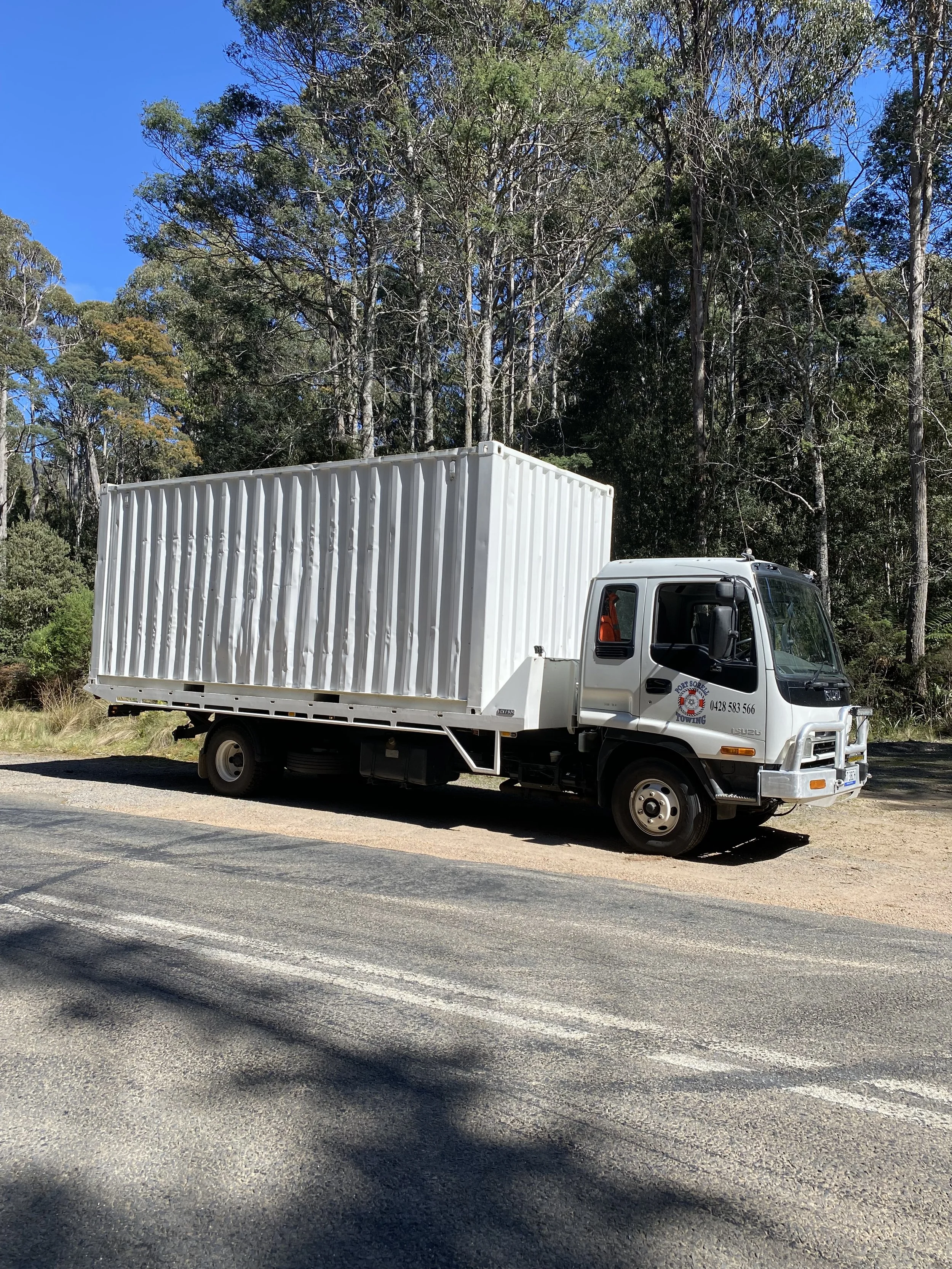 A white flatbed truck parked on the side of a rural road with a dense forest and blue sky in the background.