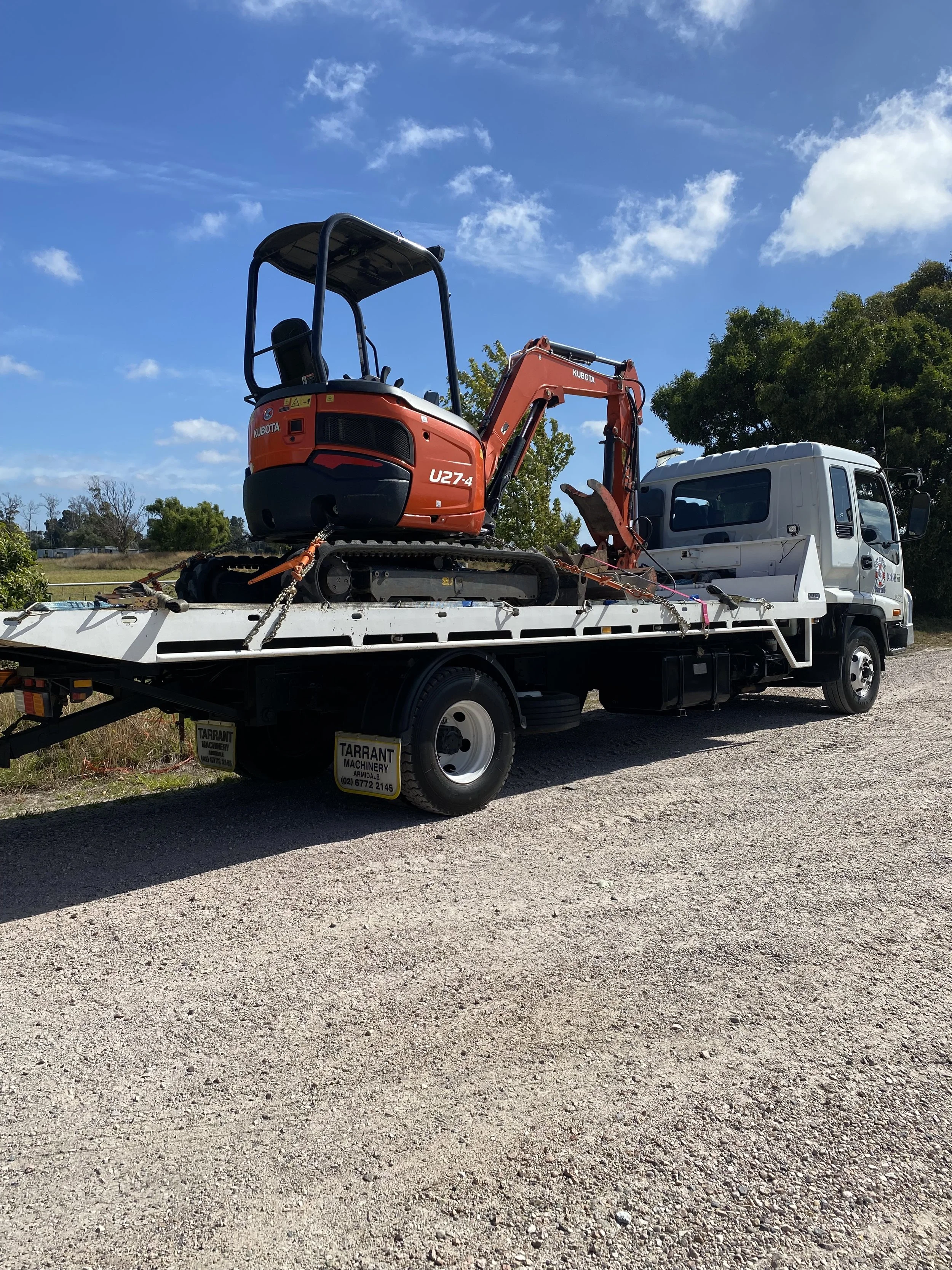A flatbed truck with a small orange excavator on its bed, parked on a gravel road with trees and a blue sky in the background.