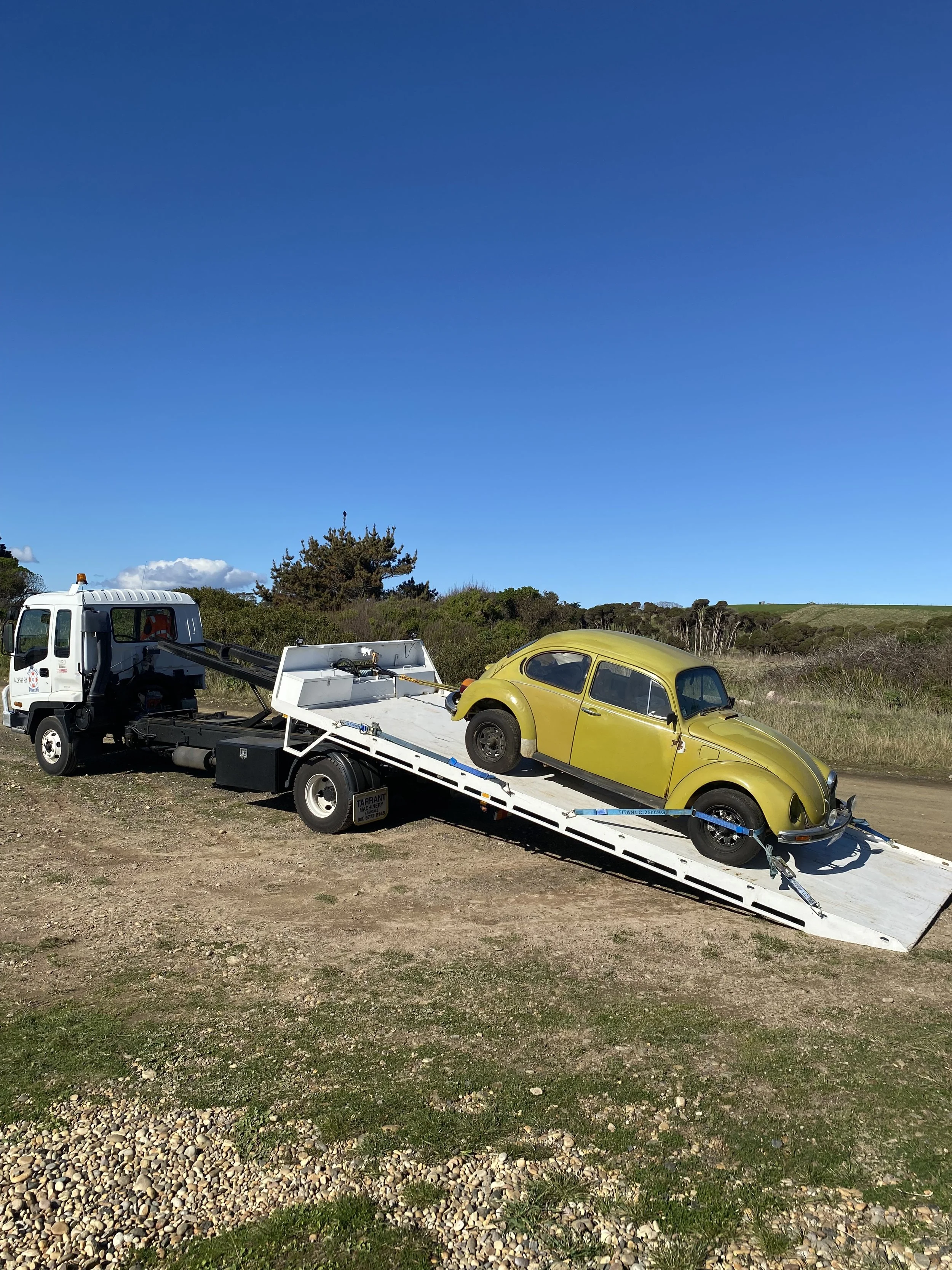 A yellow vintage car loaded on a flatbed tow truck parked on a dirt road under a blue sky.