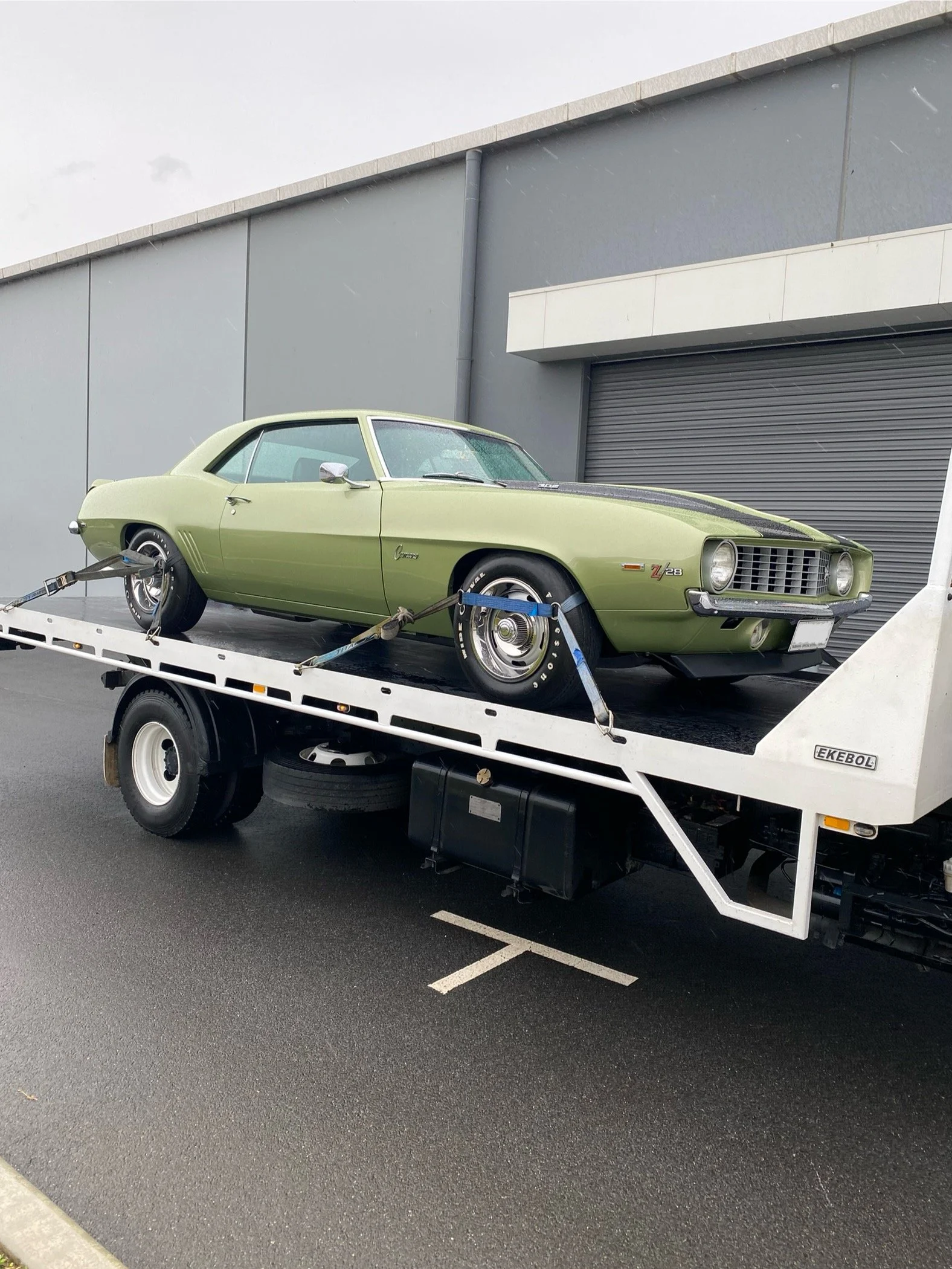 A vintage green Chevrolet Camaro Z/28 on a white car transporter truck, parked outside a modern industrial building with gray walls and a roller door.