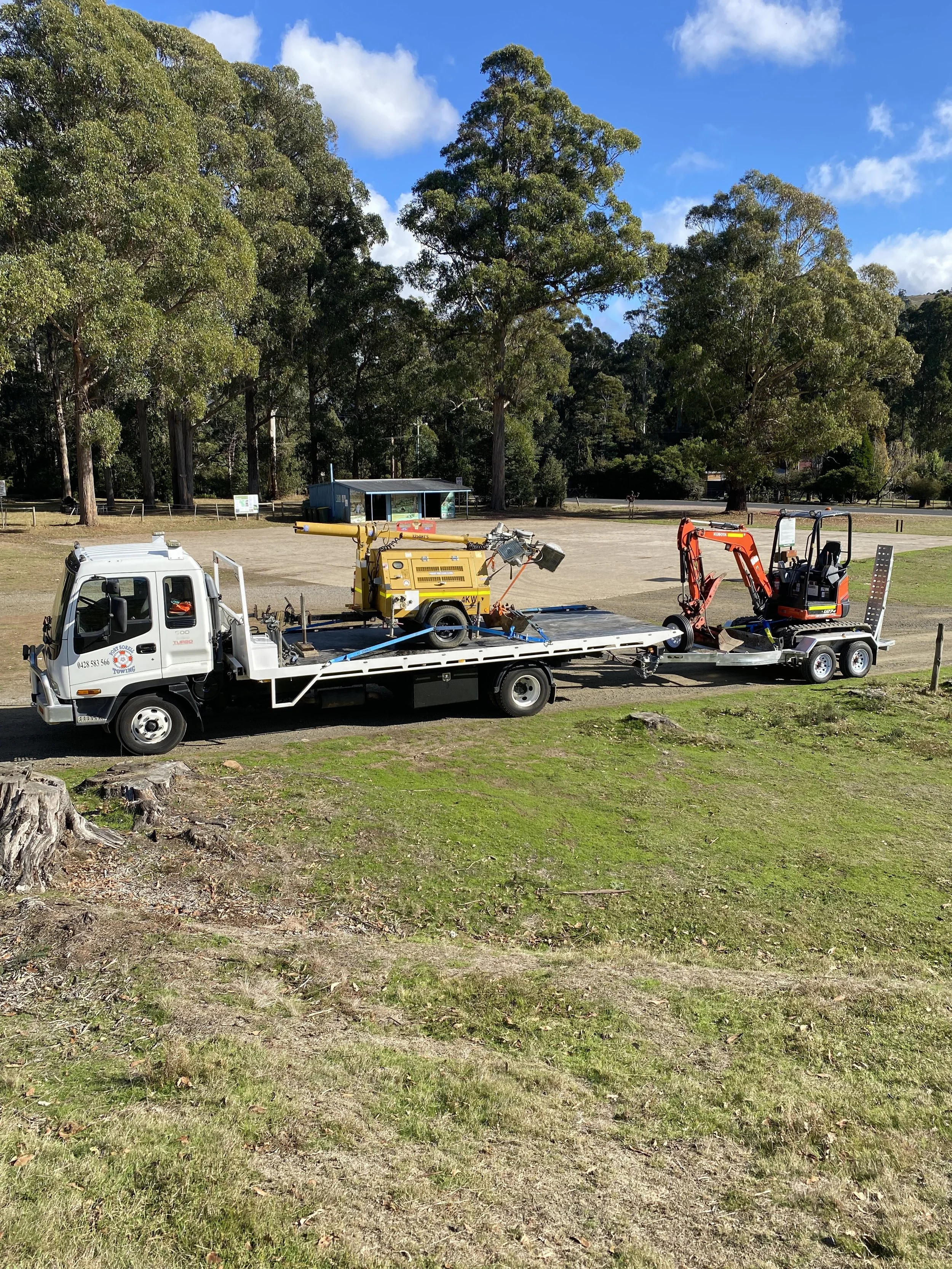 A flatbed tow truck carrying a yellow generator and a small red excavator parked on a grassy area near trees in a park.