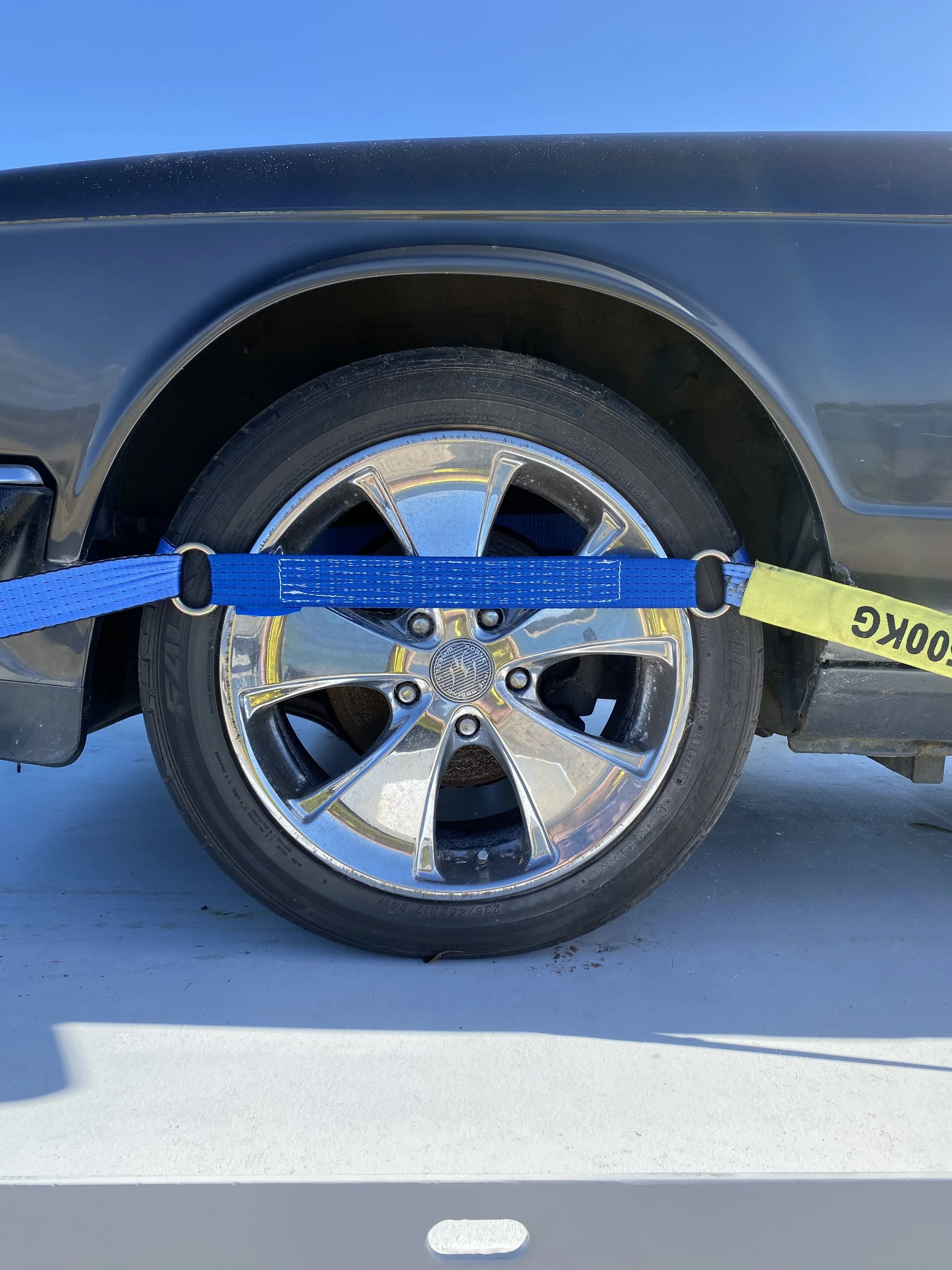 A car wheel with a chrome rim is secured with a blue strap and a yellow strap labeled 'LOCK' on a white surface against a clear blue sky.